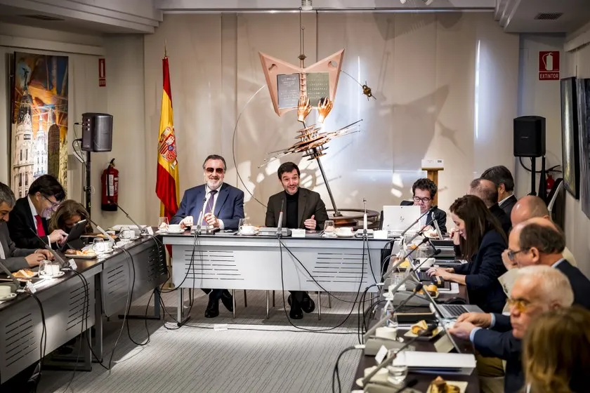 Personas sentadas en una mesa larga con micrófonos durante una conferencia de prensa en una sala con una maqueta de barco colgada del techo y una bandera española en la pared.