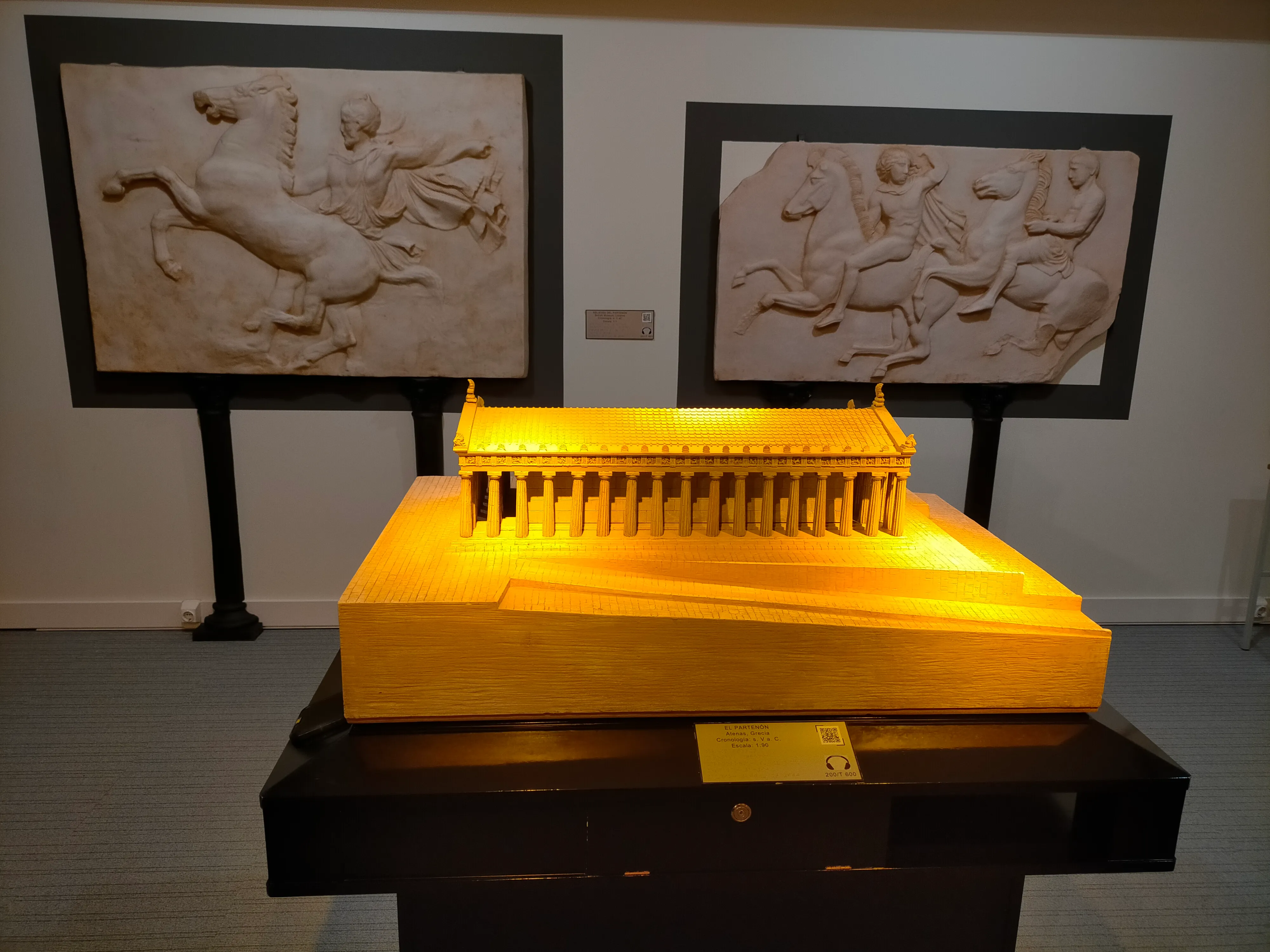 Modelo táctil del Partenón sobre un pedestal negro, iluminado en amarillo, con dos esculturas en bajorrelieve en la pared detrás.