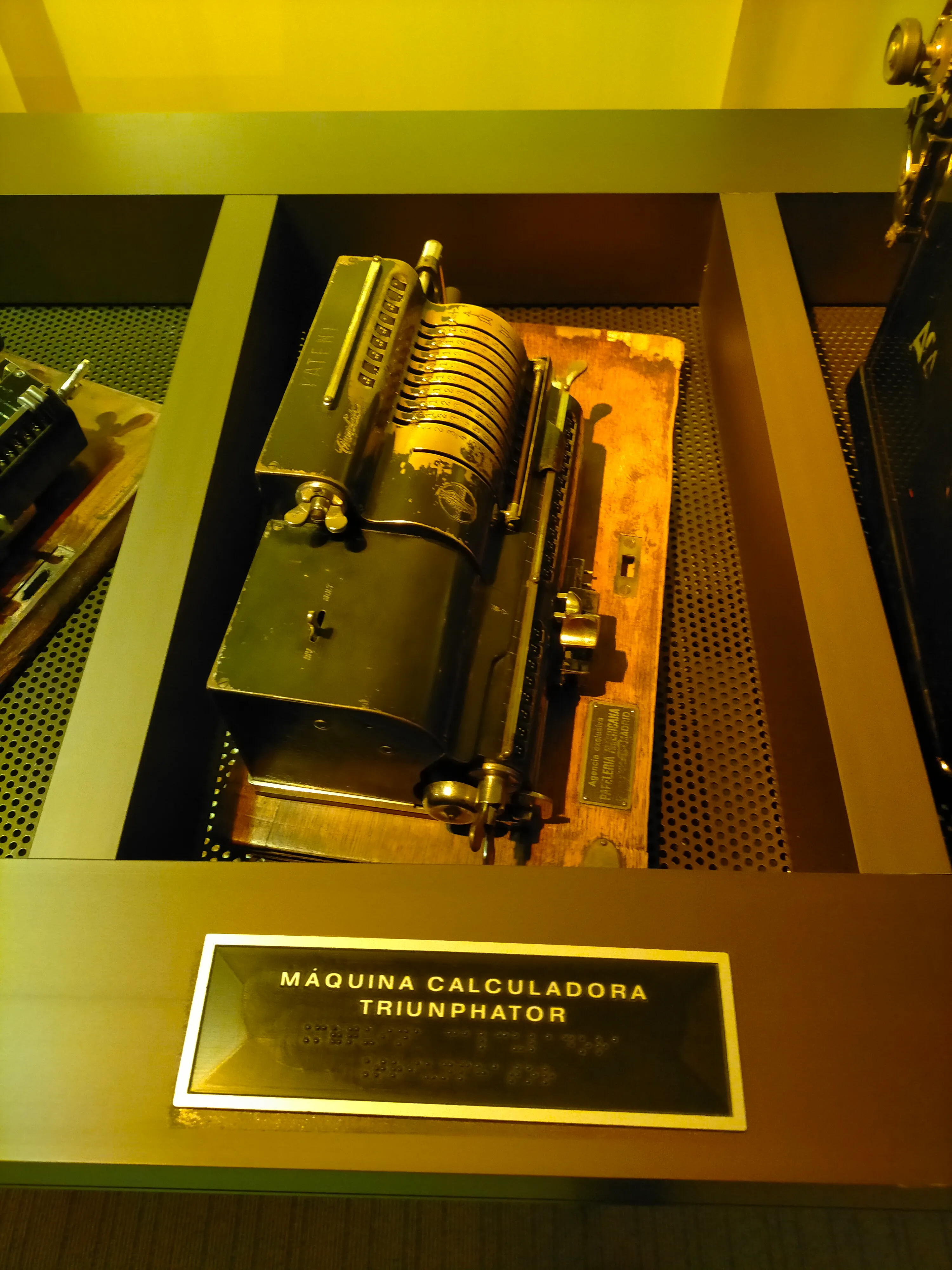 Una calculadora antigua etiquetada como 'Máquina Calculadora Triumphator' expuesta en una vitrina de museo