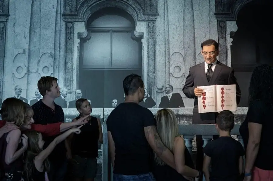 Figurka animatroniczna w garniturze trzymająca dokument, otoczona przez zwiedzających na wystawie muzealnej z klasycznym tłem architektonicznym.