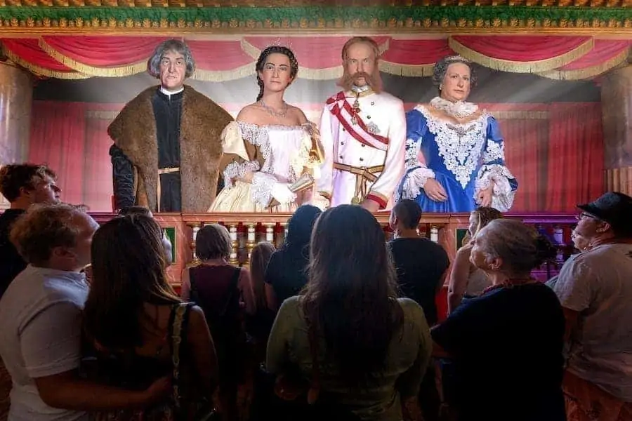 Grupa zwiedzających ogląda wystawę animatroniczną z czterema postaciami historycznymi w wyszukanych kostiumach na scenie z czerwonymi zasłonami.