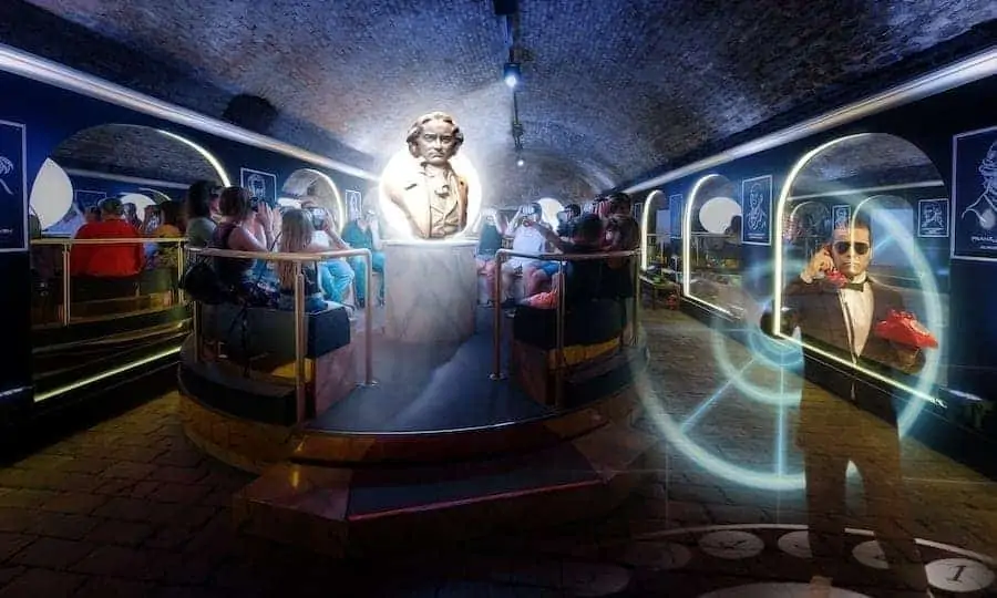 Inside a dark, tunnel-like ride with guests seated in vehicles, illuminated by blue and white lights, featuring a central holographic figure and animated characters on screens.
