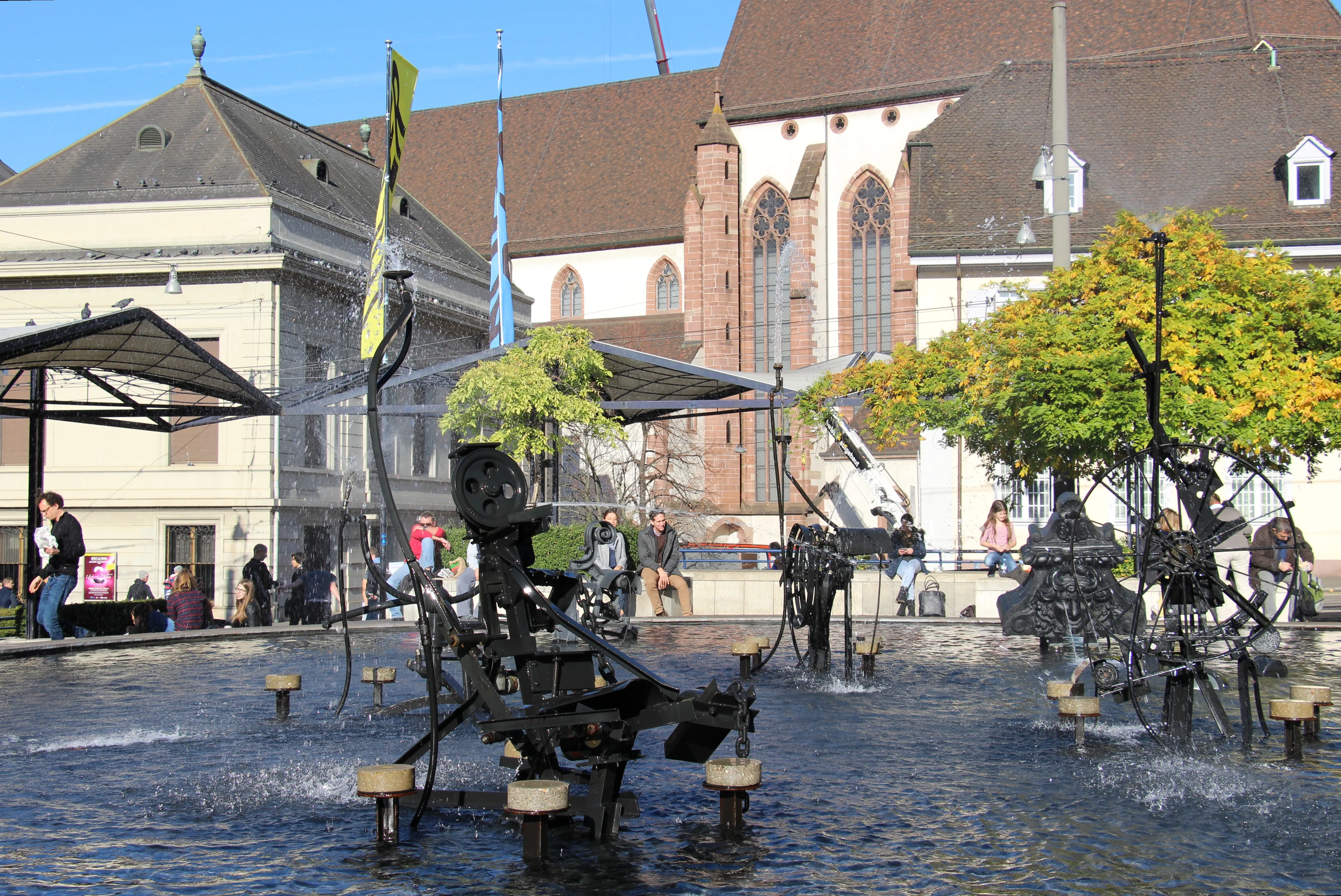 Czarne mechaniczne rzeźby kinetyczne fontanny Tinguely tryskające wodą w płytkim basenie, z historycznymi budynkami i wieżą kościoła w tle