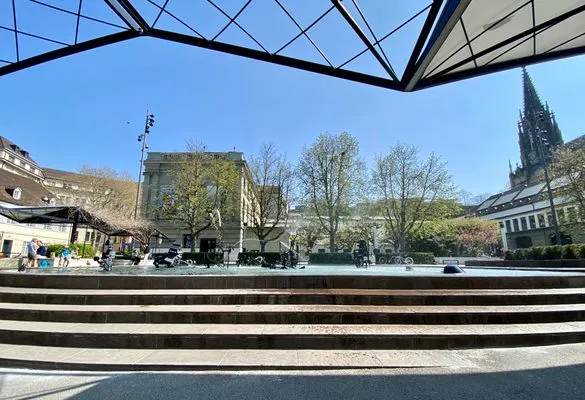 Rozległy widok na plac z fontanną Tinguely-Brunnen, ukazujący szerokie kamienne schody prowadzące do basenu z wodą, w otoczeniu budynków i wieży kościoła na tle błękitnego nieba.