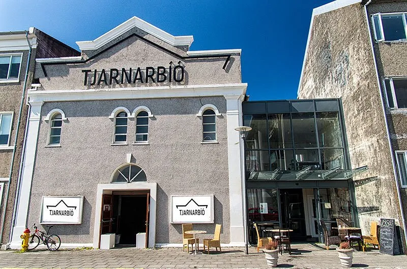 Buitenaanzicht van het Tjarnarbíó-gebouw met een gevel van grijze steen, boogvensters, de hoofdingang, terrasmeubilair en het opschrift TJARNARBÍÓ onder een helderblauwe lucht.
