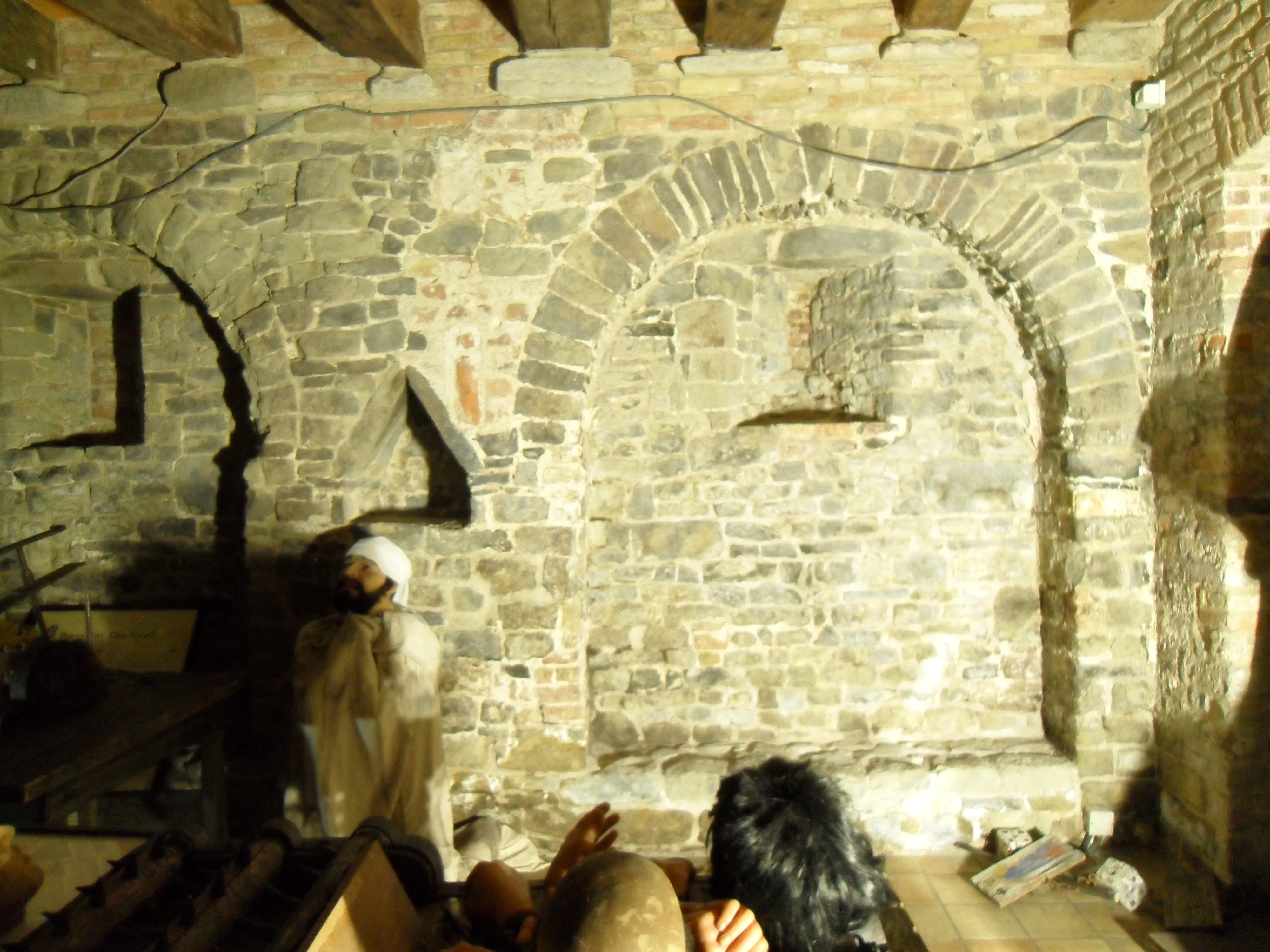 Interior de bodega de piedra medieval con nichos de arco de ladrillo, vigas de techo de madera y figuras de cera que representan una escena de mazmorra