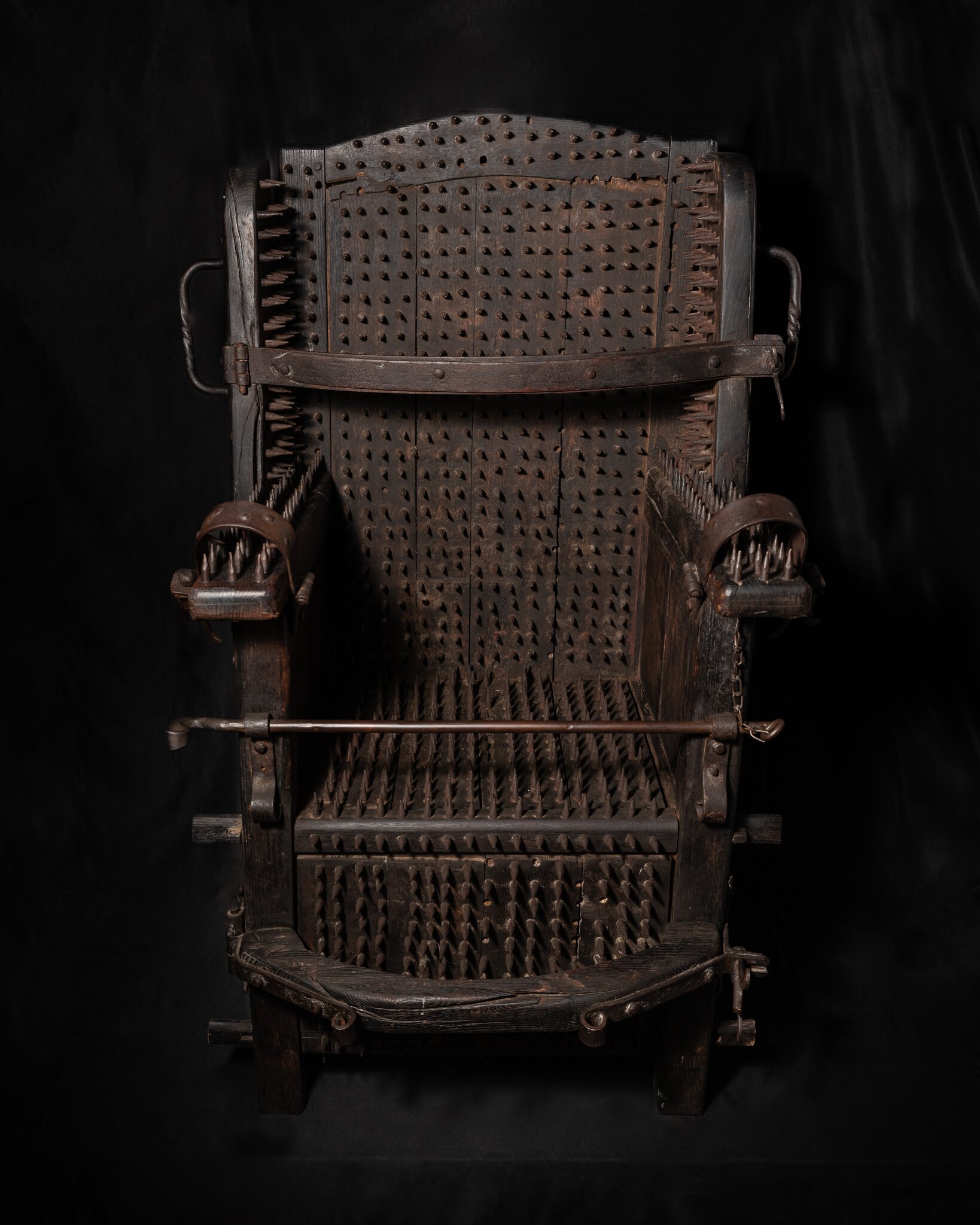 Silla de tortura de madera oscura con púas metálicas cubriendo el asiento, el respaldo y los reposabrazos sobre un fondo negro