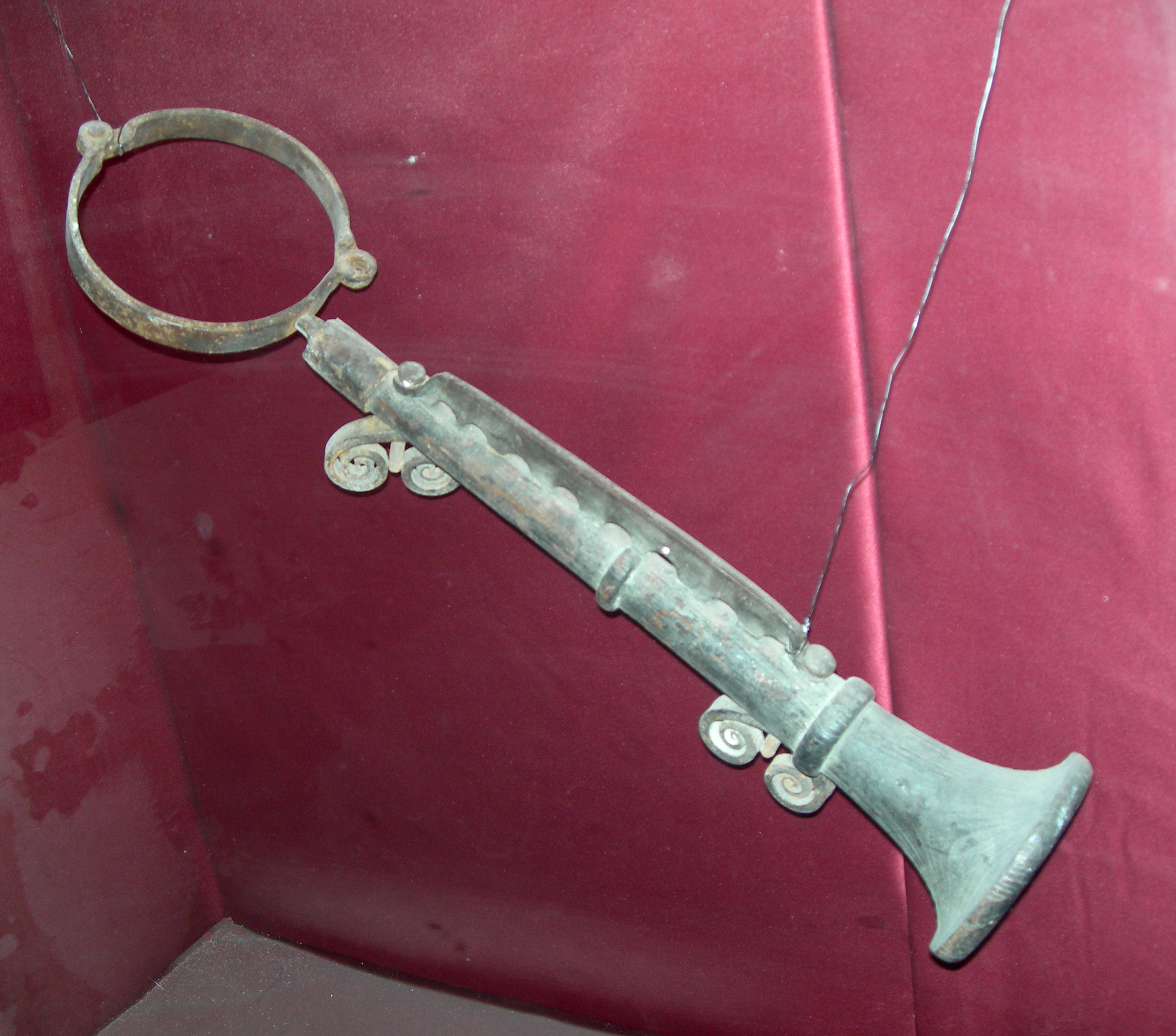 Un instrumento metálico con un anillo circular y un eje largo, expuesto sobre un fondo rojo en una vitrina de cristal