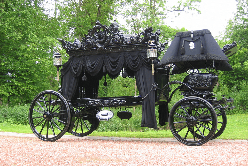 Ornate black horse-drawn funeral carriage with decorative metalwork and draped fabric, displayed outdoors against a green tree backdrop at the museum
