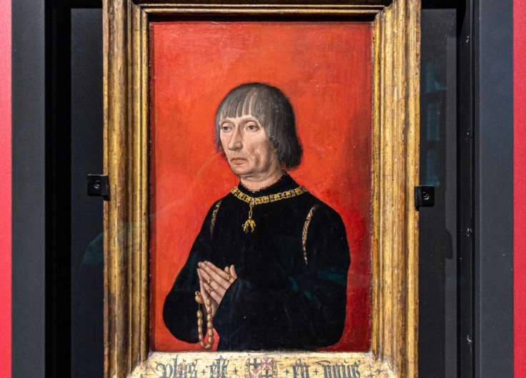 Retrato histórico enmarcado de una persona vestida de negro con ribete dorado sobre fondo rojo, expuesto en vitrina de museo