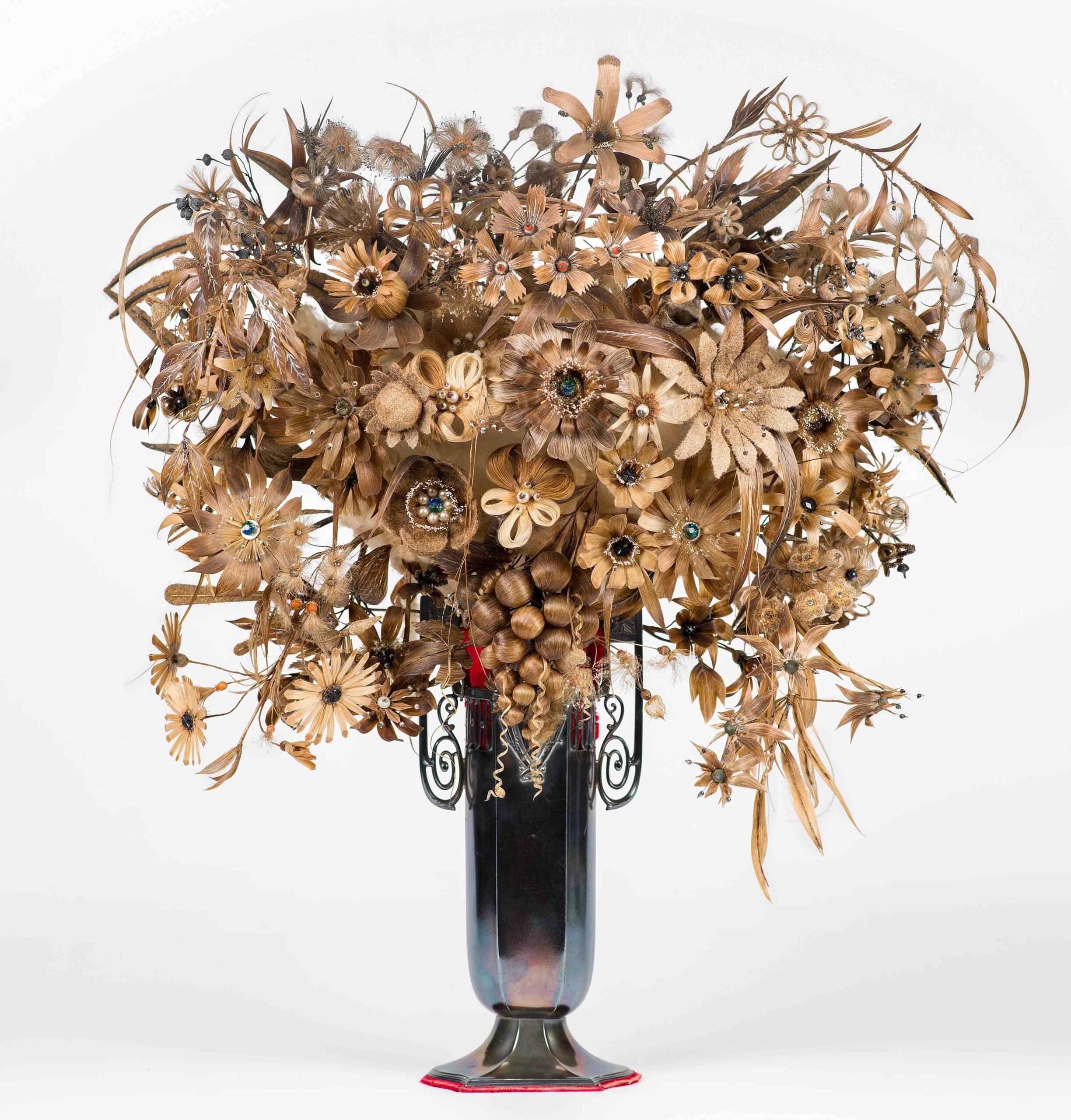 Arreglo floral seco y marrón con base intrincada de metal, que presenta diversos materiales vegetales conservados en un ornamentado jarrón oscuro