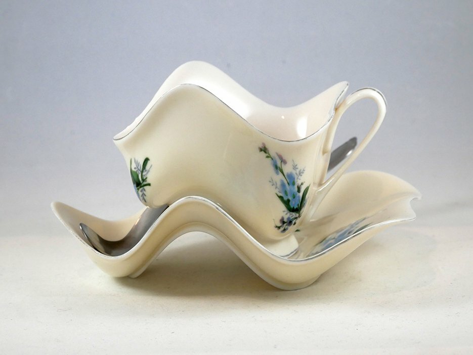 Taza y platillo de cerámica blanca con motivos florales azules sobre una base ondulada, expuestos sobre un fondo claro