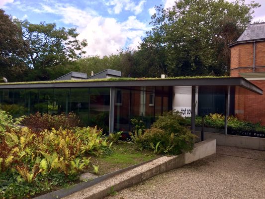 Modern building with green roof and glass facade, surrounded by garden plants, sign reading 'tot zo' visible near entrance