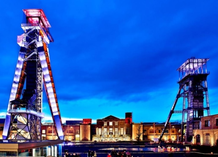 Twilight exterior view of a building with two illuminated lattice tower structures, blue sky, and reflecting water