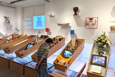 Museum interior with casket-shaped display cases containing digital screens, a floral arrangement, and a visitor examining funeral culture exhibits.
