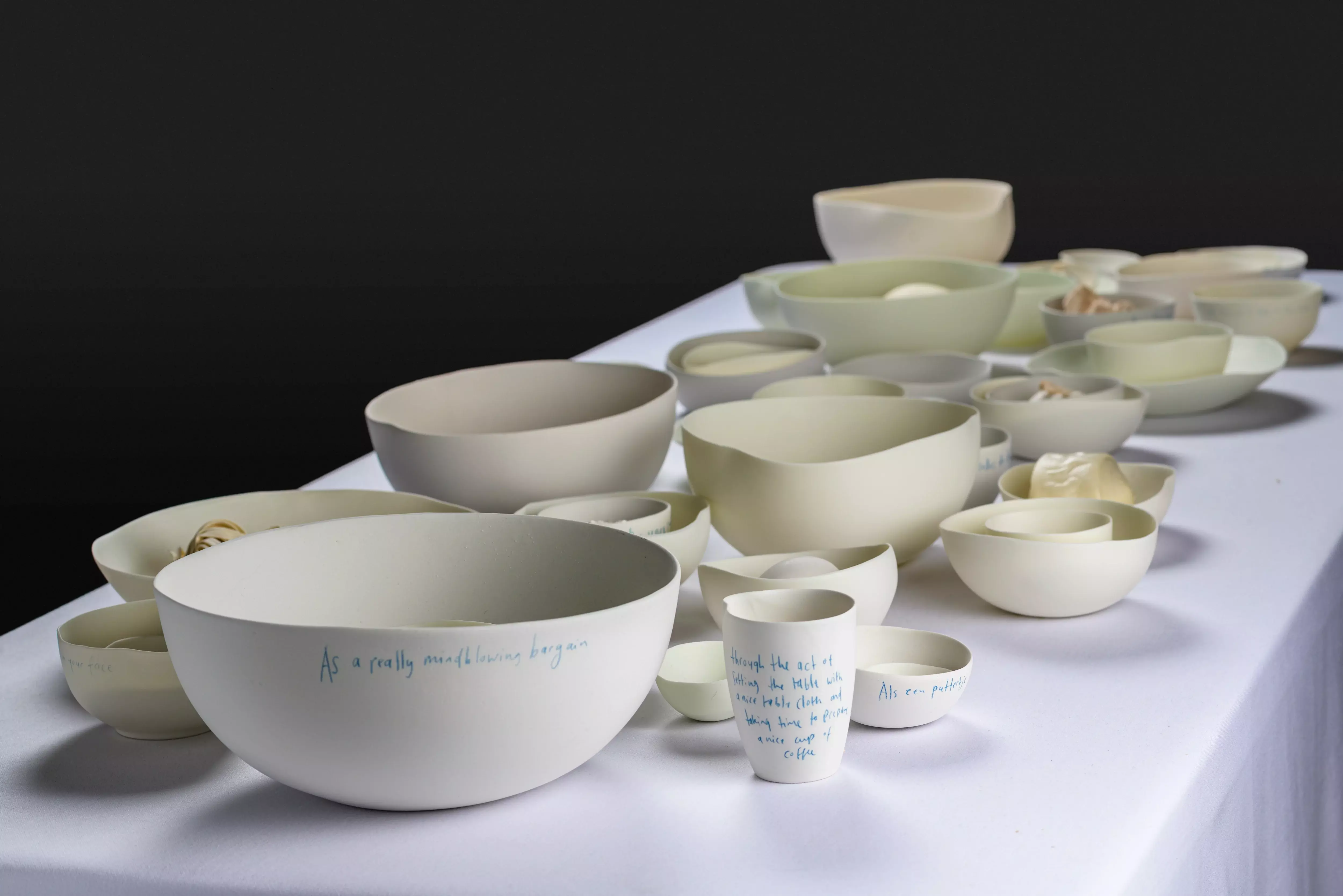 Cuencos y tazas de cerámica blanca con texto escrito a mano expuestos sobre una mesa blanca contra un fondo oscuro, parte de una exposición sobre la cultura funeraria.