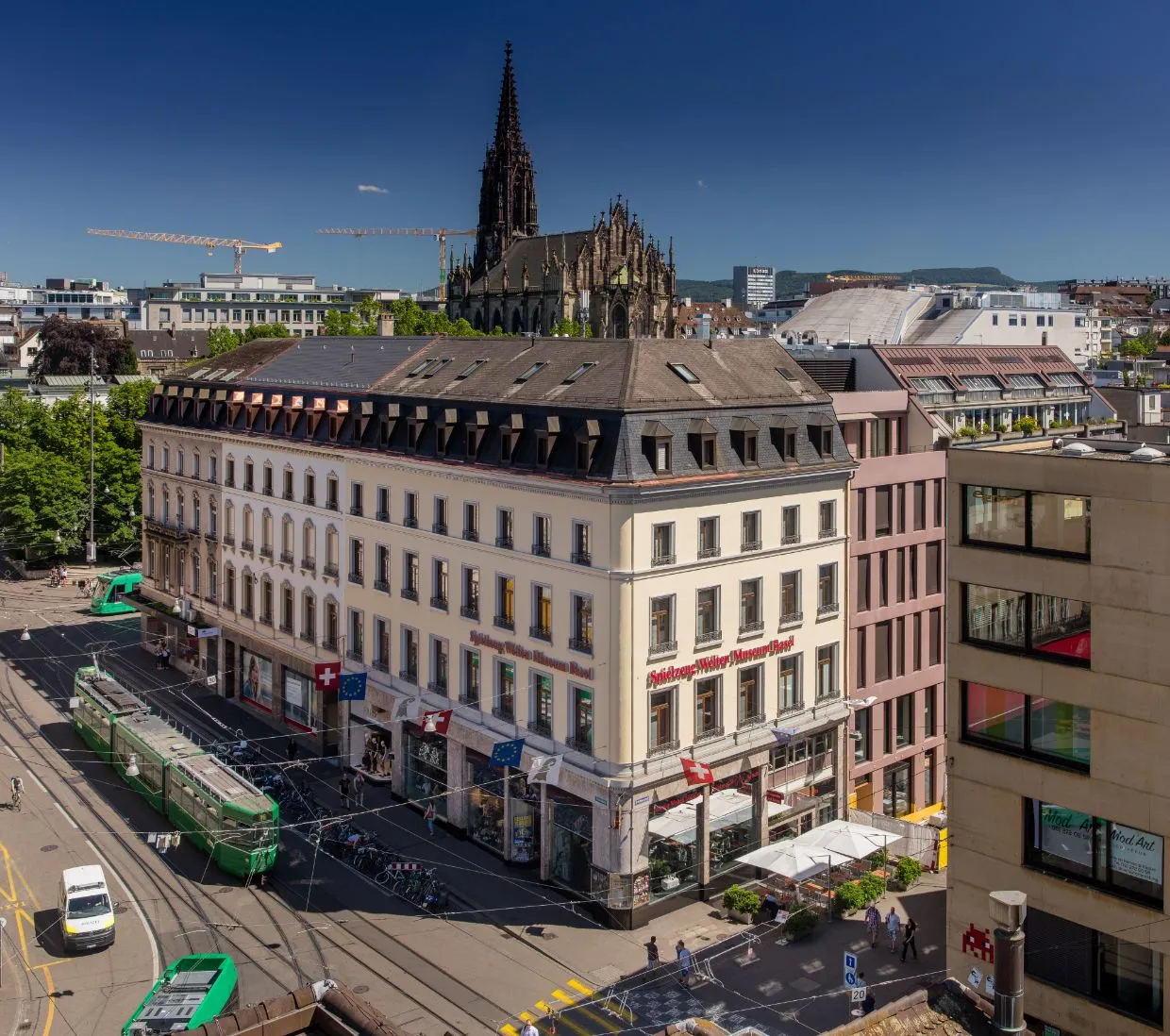 Luchtfoto van een meerverdiepingsgebouw op een straathoek in Bazel met een groene tram die voorbijrijdt en een gotische kerktoren die op de achtergrond uitsteekt onder een helderblauwe lucht.