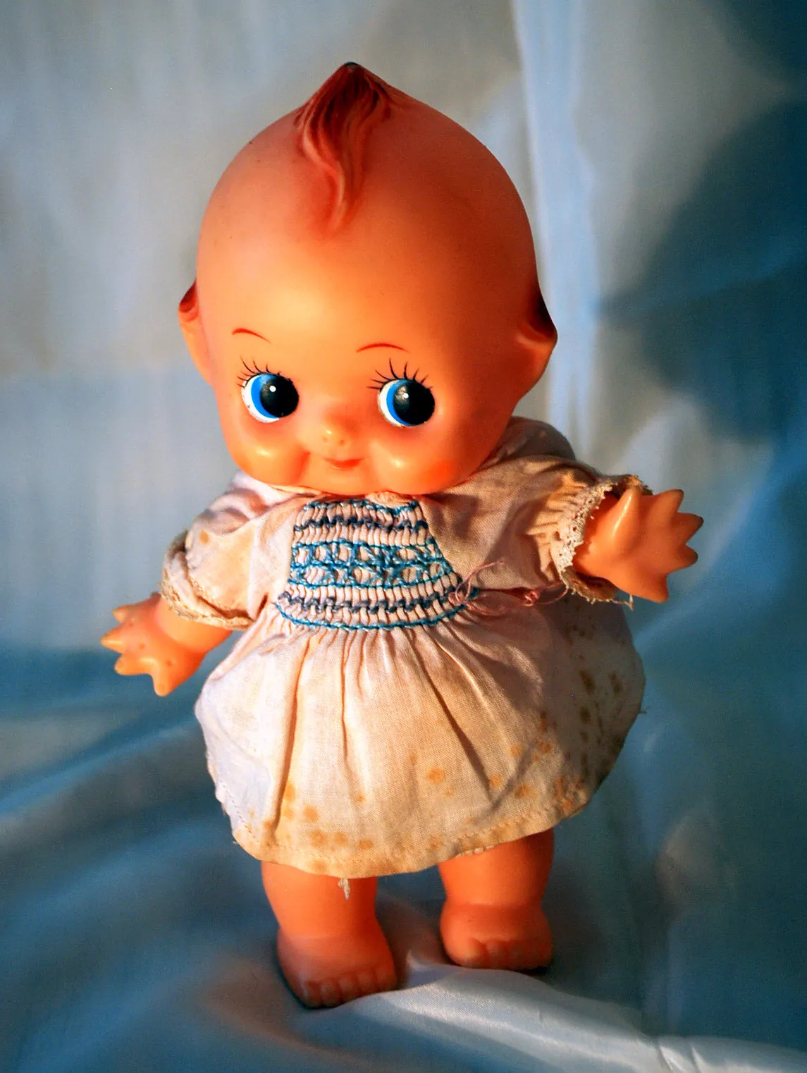 Een vintage Kewpie-pop met grote blauwe ogen, een plukje roodachtig haar en een witte smockjurk, staand tegen een lichtblauwe stoffen achtergrond