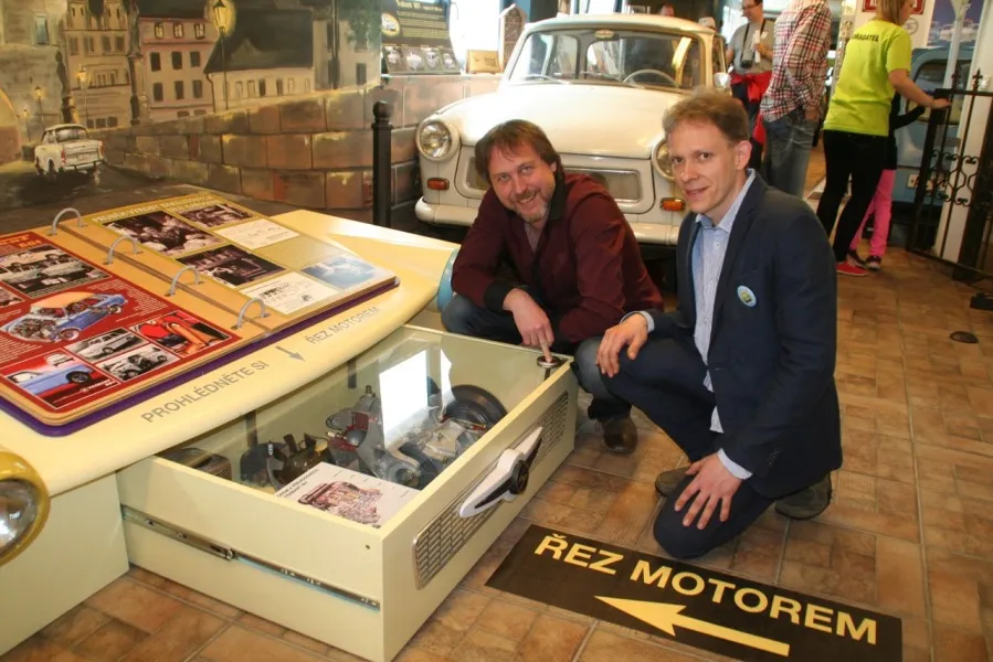 Dos hombres agachados junto a una vitrina con recuerdos del Trabant y un coche Trabant blanco al fondo.