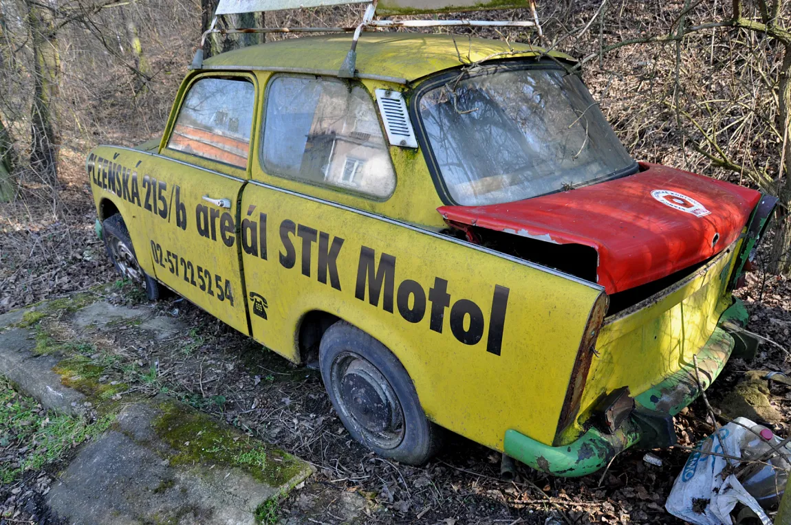 Żółty samochód Trabant z czerwonym dachem zaparkowany na zewnątrz, z napisem 'STK Motol' z boku.