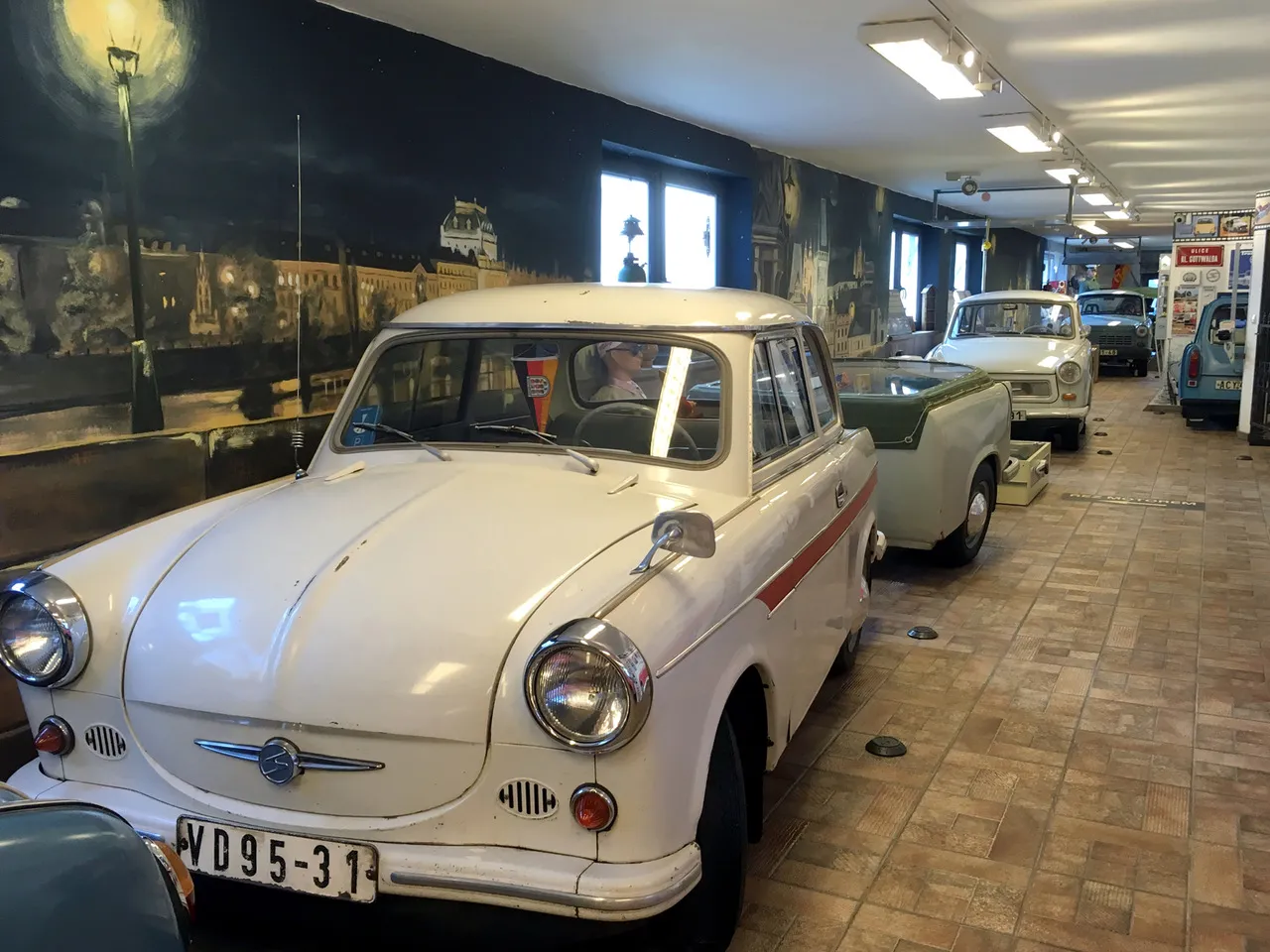 Biały samochód Trabant z czerwoną paskiem wyeksponowany w muzeum wśród innych zabytkowych samochodów i muralu na ścianie
