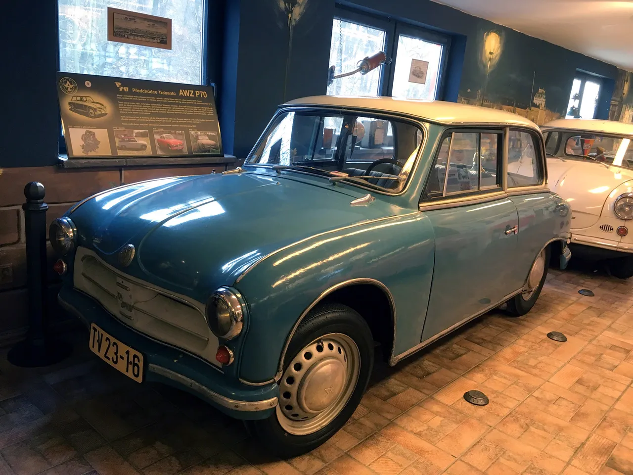 Jasnoniebieski, zabytkowy samochód Trabant z białym dachem, wystawiony wewnątrz na wyłożonej kafelkami podłodze, z tablicą rejestracyjną o numerze 'IV 23-16'.
