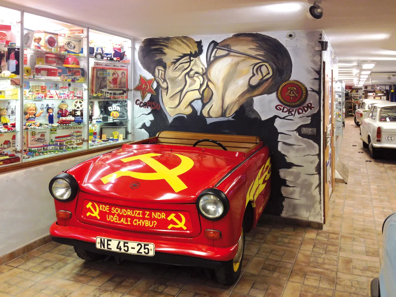 Czerwony samochód Trabant z emblematem sierpa i młota oraz czeskim napisem na masce, wystawiony wewnątrz przed politycznym muralem.