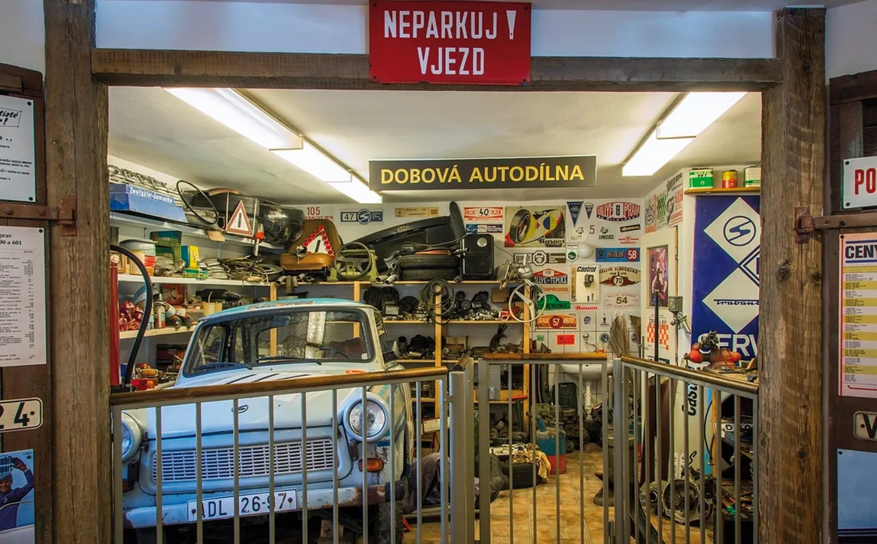 Stary niebieski samochód Trabant we wnętrzu muzeum, widziany przez drewniane drzwi z napisami 'NEPARKUJ! VJEZD' i 'DOBOVÁ AUTODÍLNA'.
