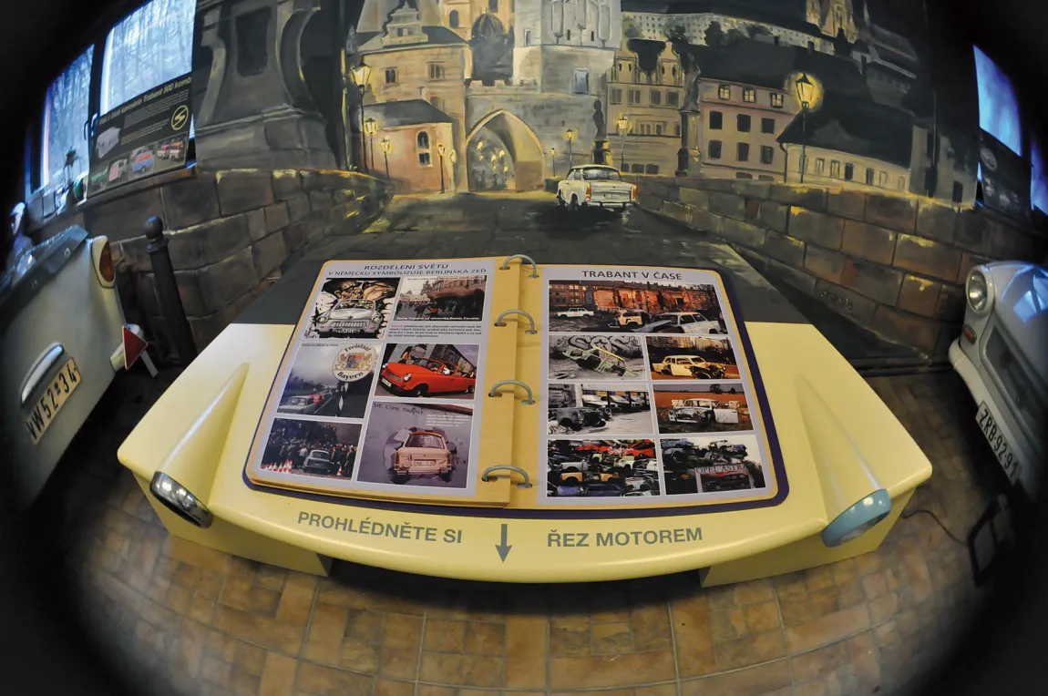 Un álbum de fotos con imágenes de coches Trabant en una pantalla amarilla con forma de coche con texto checo, sobre un telón de fondo de mural pintado.