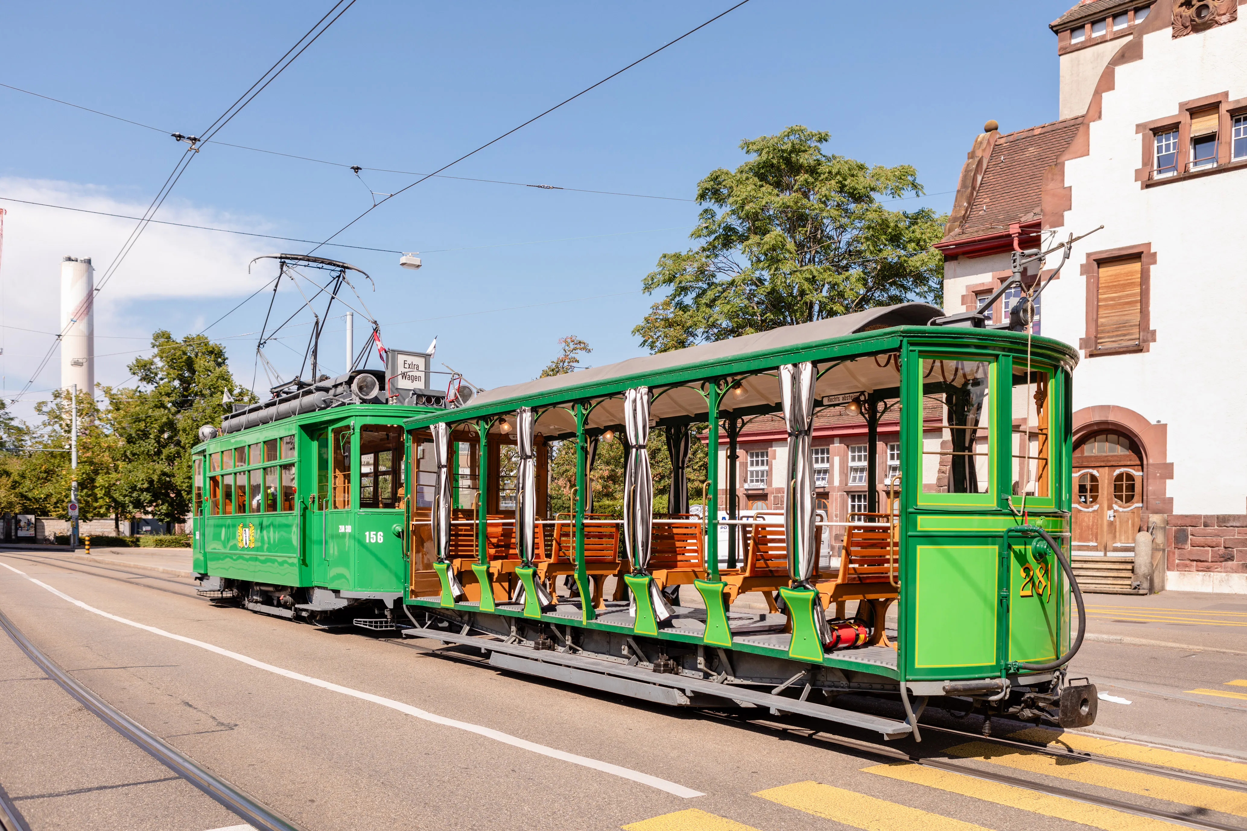 A historic green open-sided summer tram with wooden benches on tracks in front of a white building with red sandstone details in Basel.