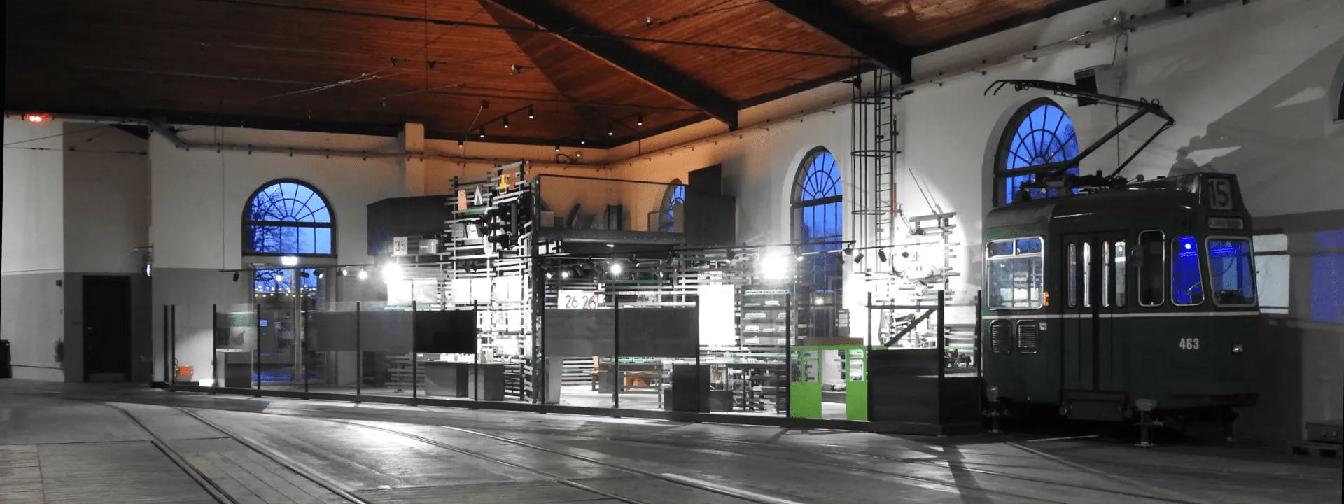 Interior of a large museum hall with historic tram vehicles on display, tall arched blue windows, exposed wooden ceiling beams, and overhead catenary equipment.