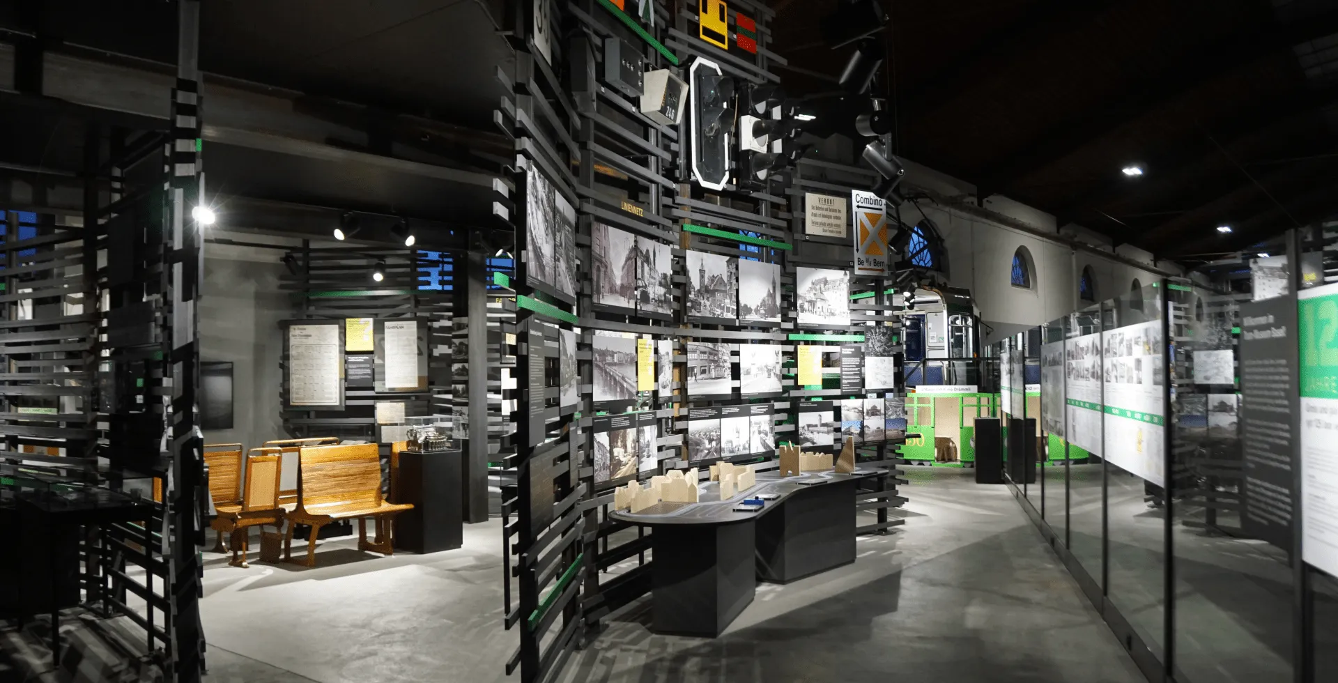 Interior of Tram-Museum Basel showing dark exhibition halls with framed photographs on walls, display cases, wooden benches, and informational panels under spot lighting