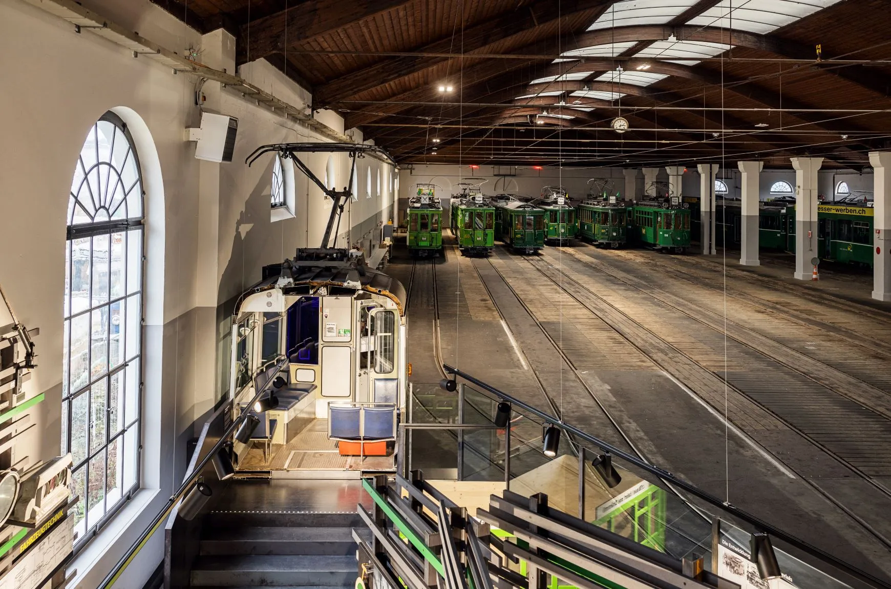 Interior of a large tram depot hall with a wooden roof, arched windows, and multiple historic green trams parked on tracks, with a vintage tram near a staircase in the foreground.