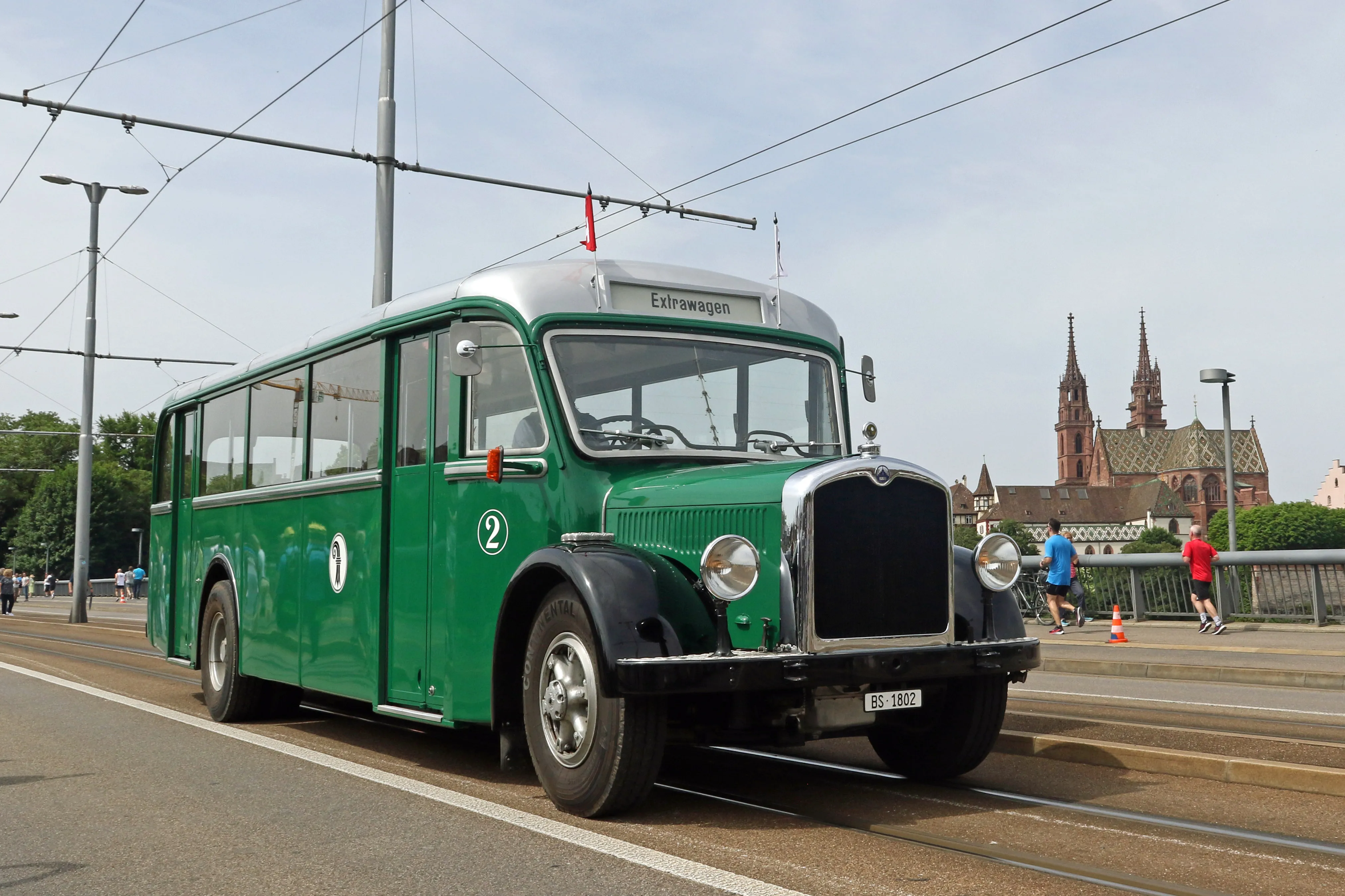Vintage green trolleybus numbered 2 with overhead wires on a bridge, with Basel Minster cathedral spires visible in the background