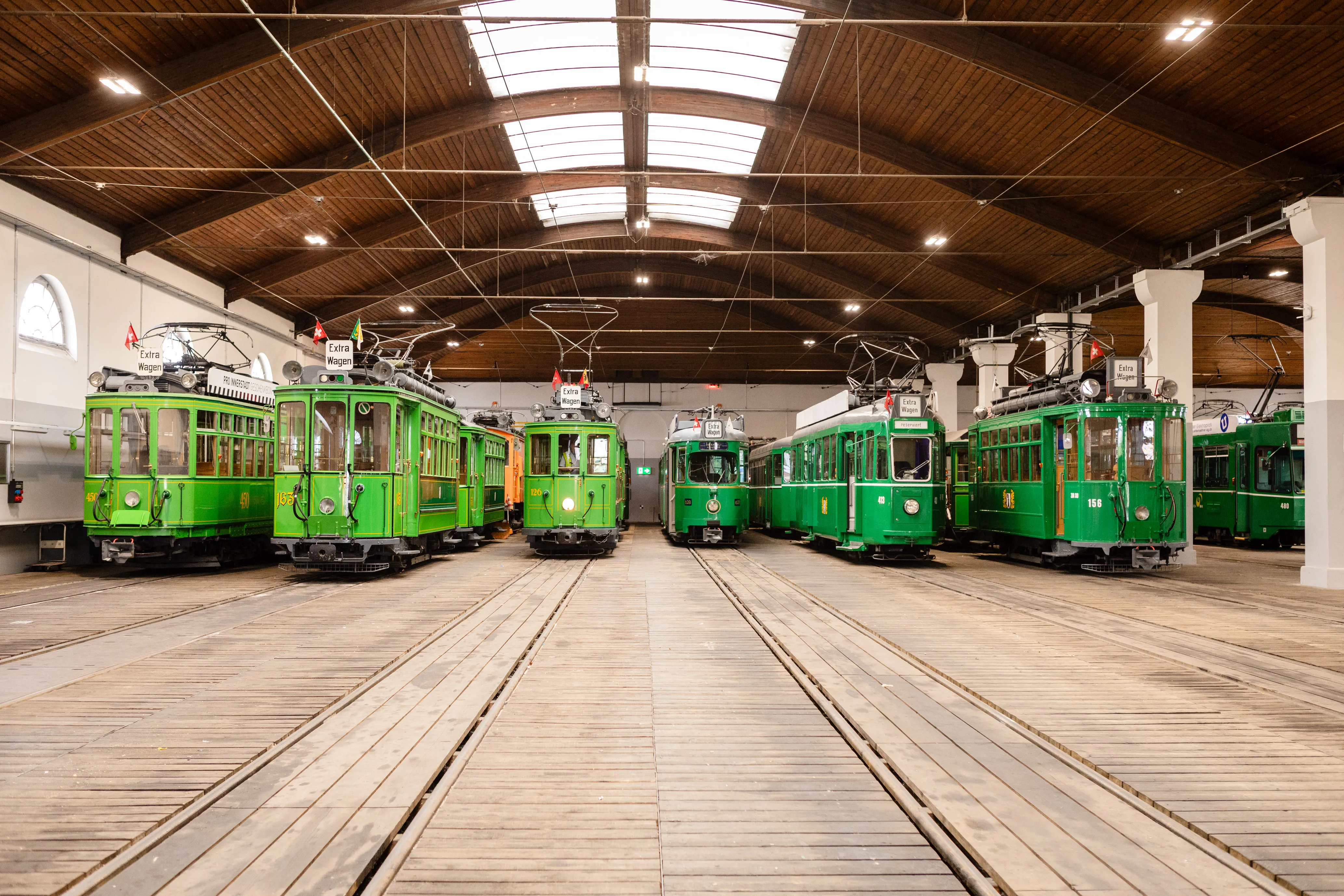 Rows of vintage green trams parked on parallel tracks inside a large wooden-roofed hall with skylights at the Tram-Museum Basel