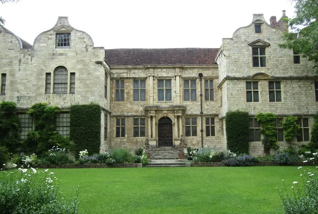Stone facade of a historic house with symmetrical gabled wings, central entrance with steps, and a manicured green lawn in the foreground
