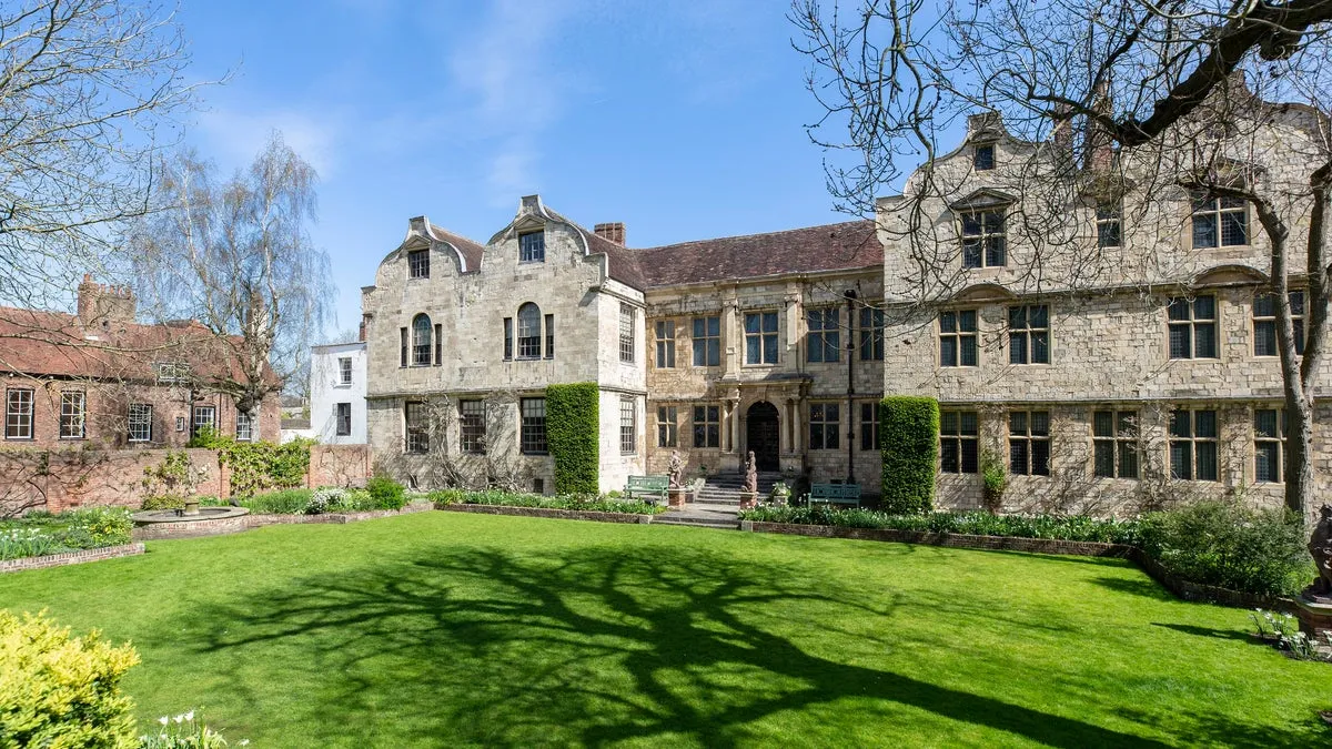 Stone historic building facade viewed from a green lawn courtyard with tree shadows, under a blue sky