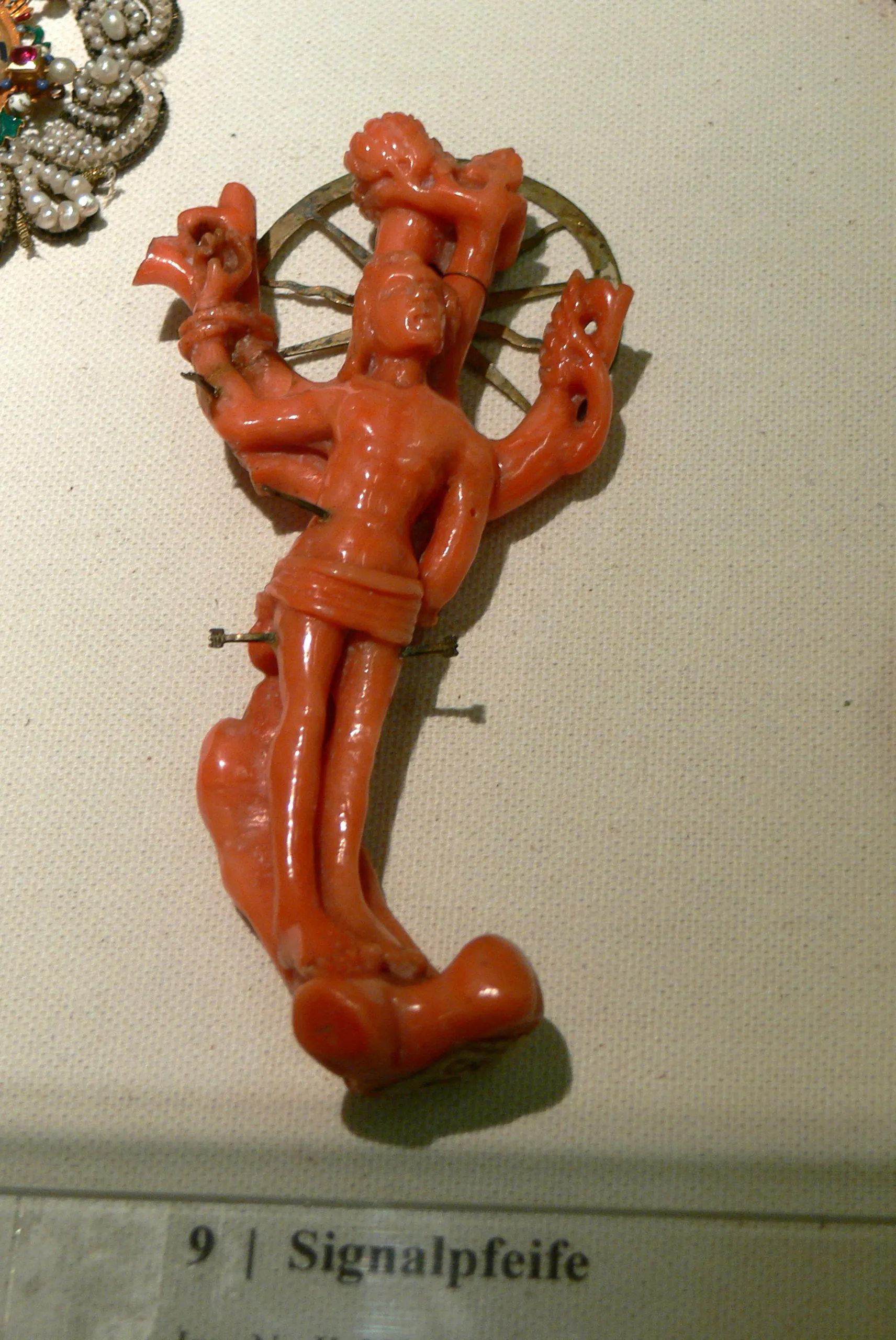 Een koraalkleurige, gesneden broche van een figuur met meerdere armen, tentoongesteld in een vitrine