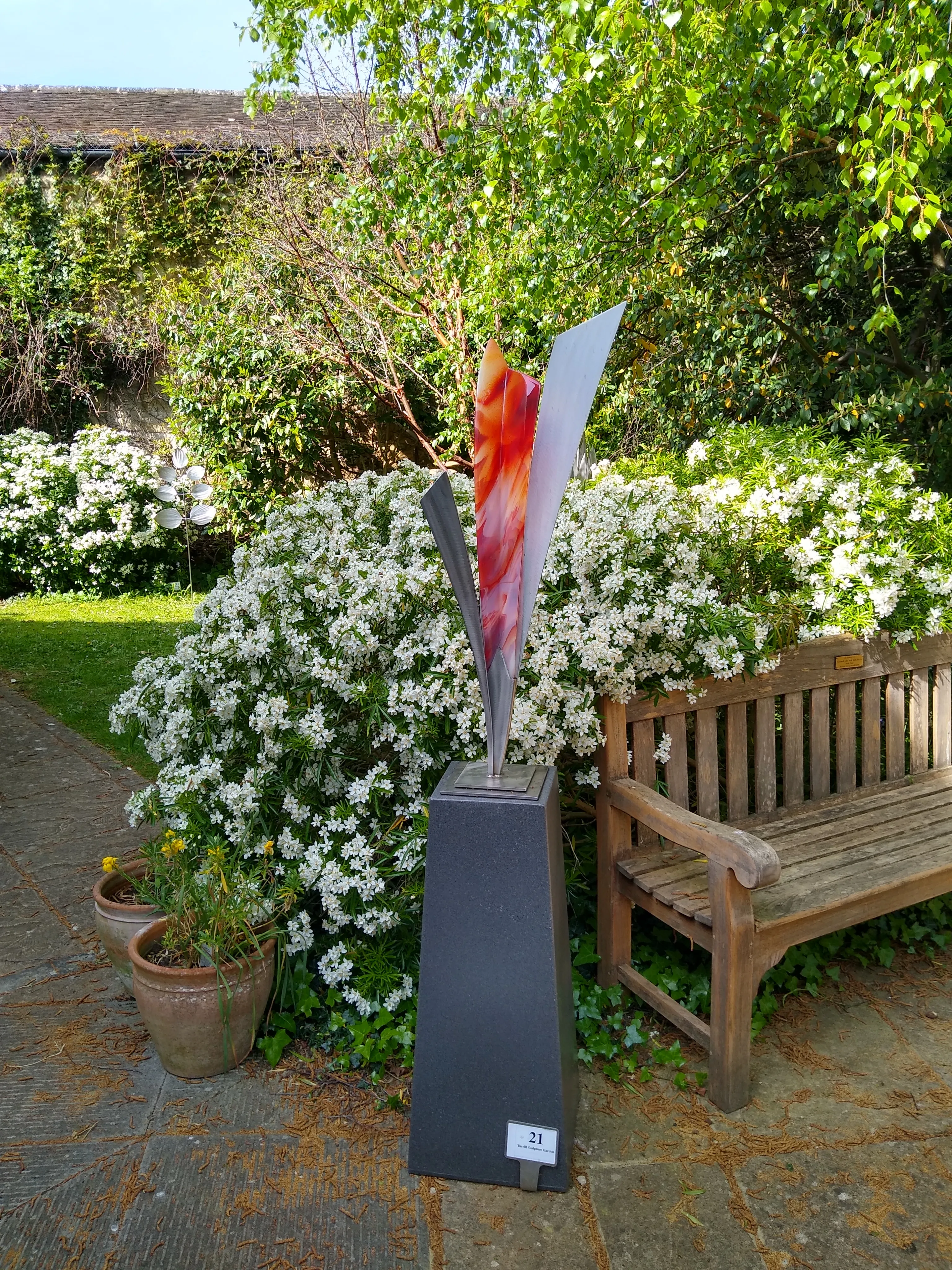 Abstracte sculptuur met rode, oranje en zilveren vormen op een sokkel in een tuin naast een houten bank en witte bloemen.