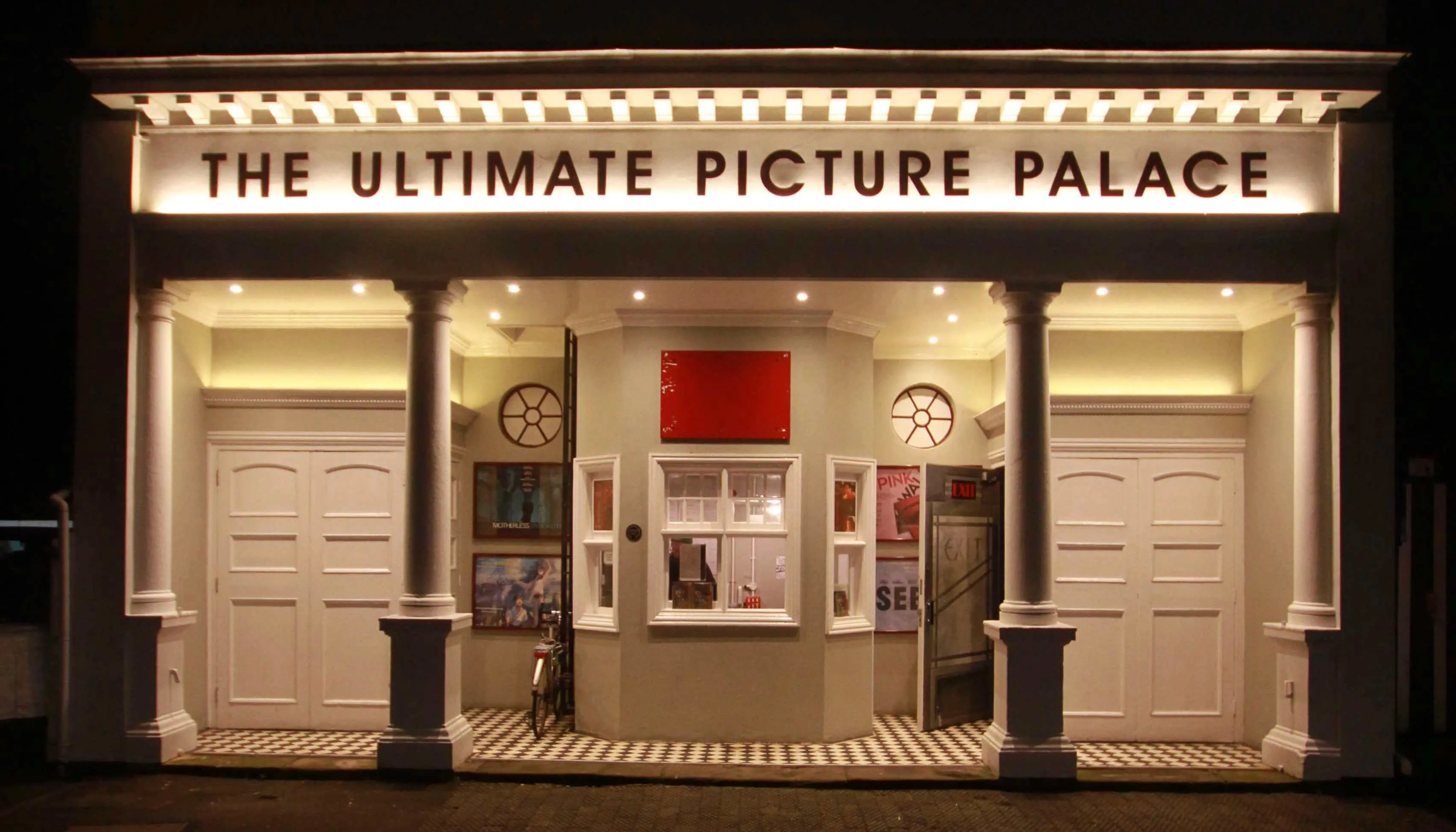 Nocny widok na fasadę kina The Ultimate Picture Palace z podświetlanym szyldem, białymi kolumnami, centralną kasą biletową z czerwonym panelem, dekoracjami w kształcie szpuli filmowej oraz plakatami filmowymi