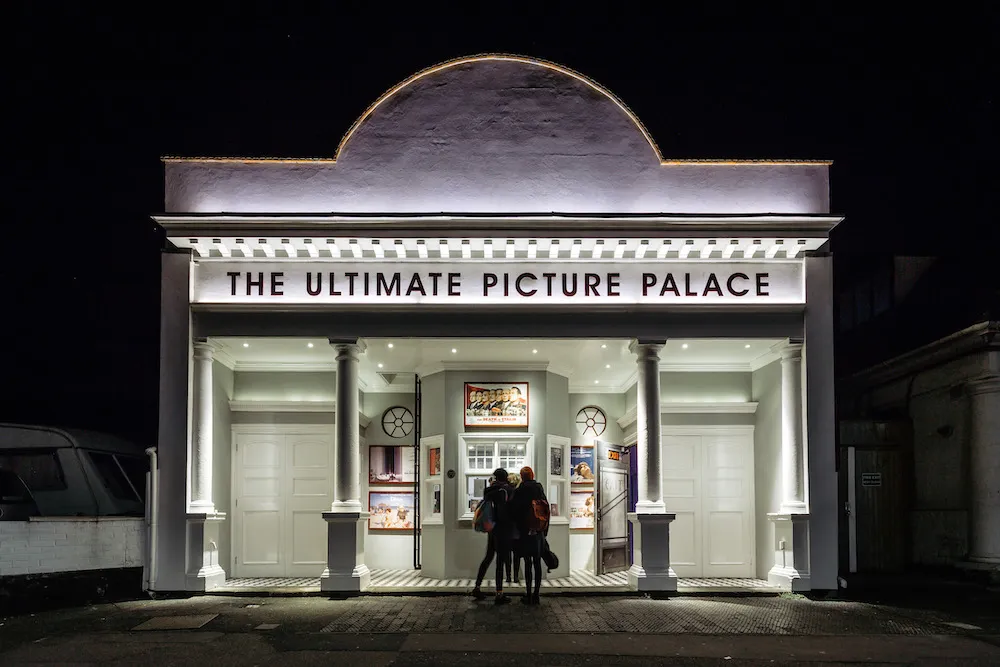 Nocny widok z zewnątrz na kino The Ultimate Picture Palace z podświetlanym szyldem z nazwą obiektu oraz dwiema osobami stojącymi przy wejściu pod łukowatą fasadą z kolumnami.
