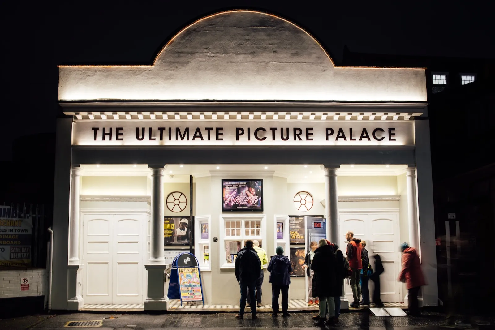 Oświetlona fasada kina The Ultimate Picture Palace w nocy, z ludźmi stojącymi na zewnątrz i czytającymi afisze filmowe