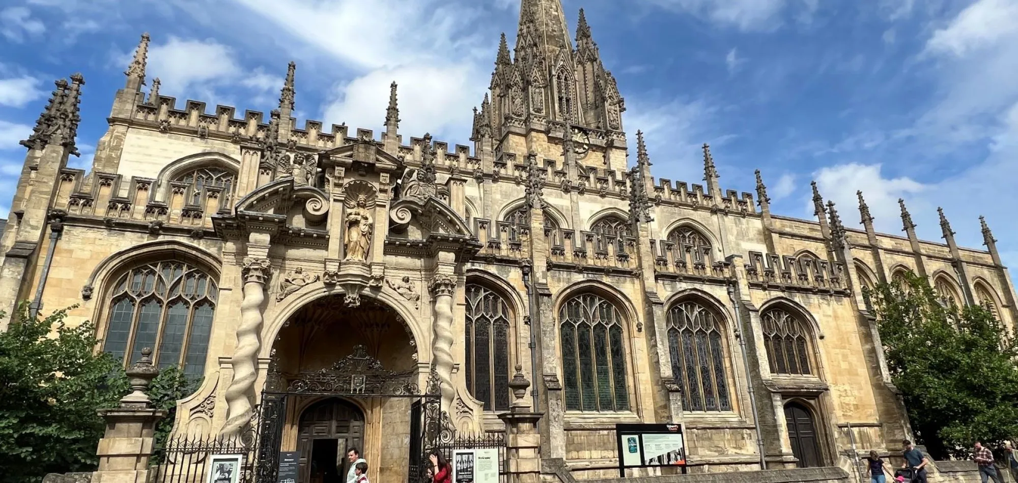 Fachada de una iglesia gótica de piedra con una alta aguja central, pináculos ornamentados, grandes ventanales arqueados y una entrada en arco con visitantes cerca