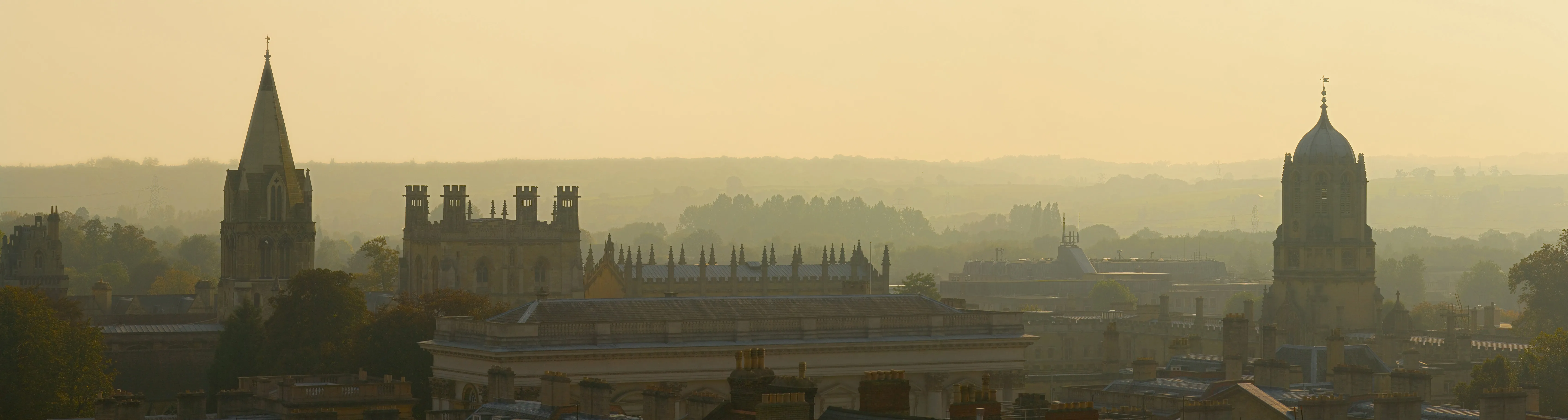 Vista panorámica del perfil urbano de Oxford bajo una neblinosa luz dorada, mostrando la aguja de la University Church of St Mary the Virgin a la izquierda y la Radcliffe Camera a la derecha.