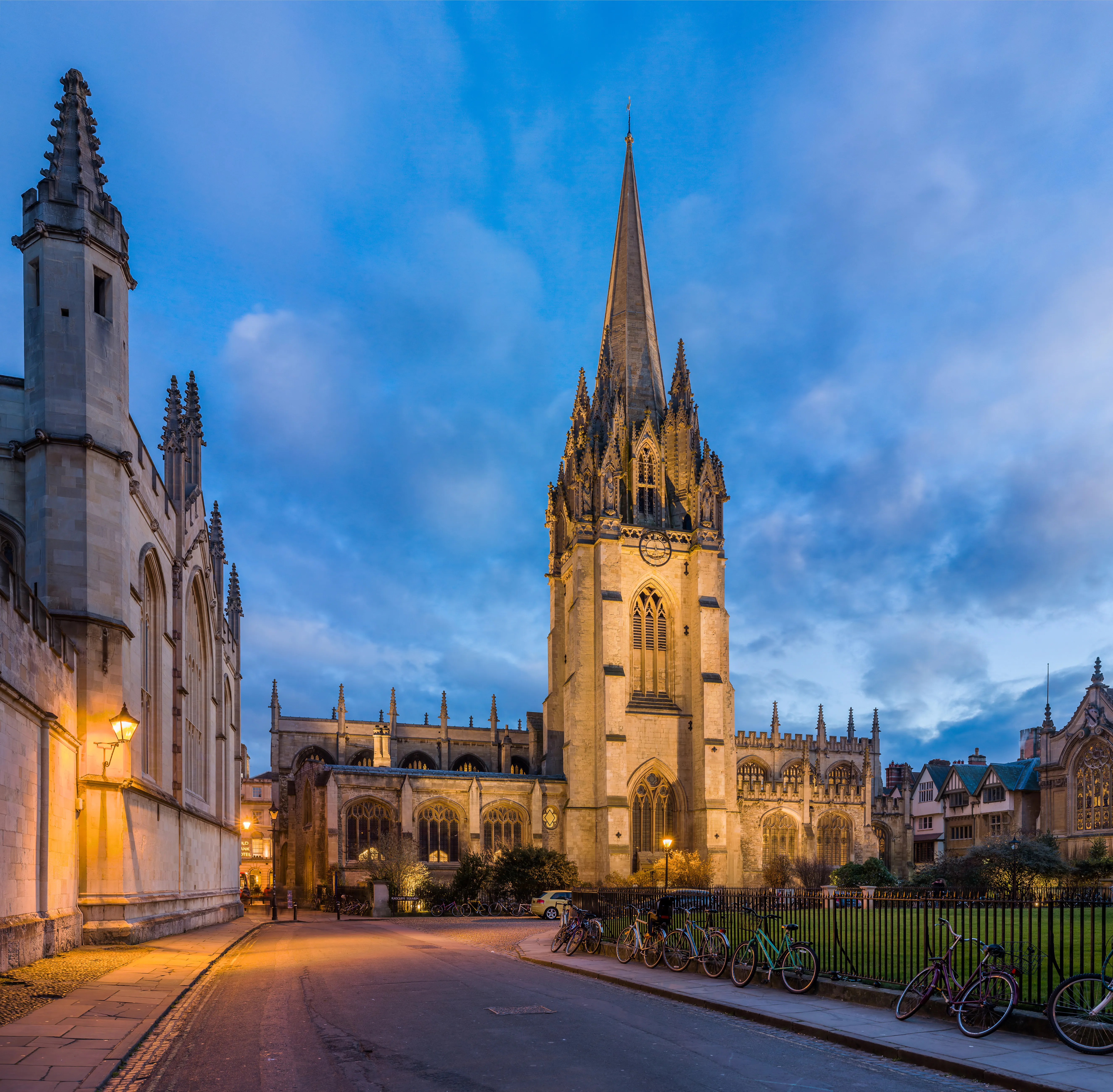 La University Church of St Mary the Virgin iluminada al atardecer, con su alta aguja gótica alzándose contra un cielo azul y bicicletas aparcadas a lo largo de una valla en primer plano.