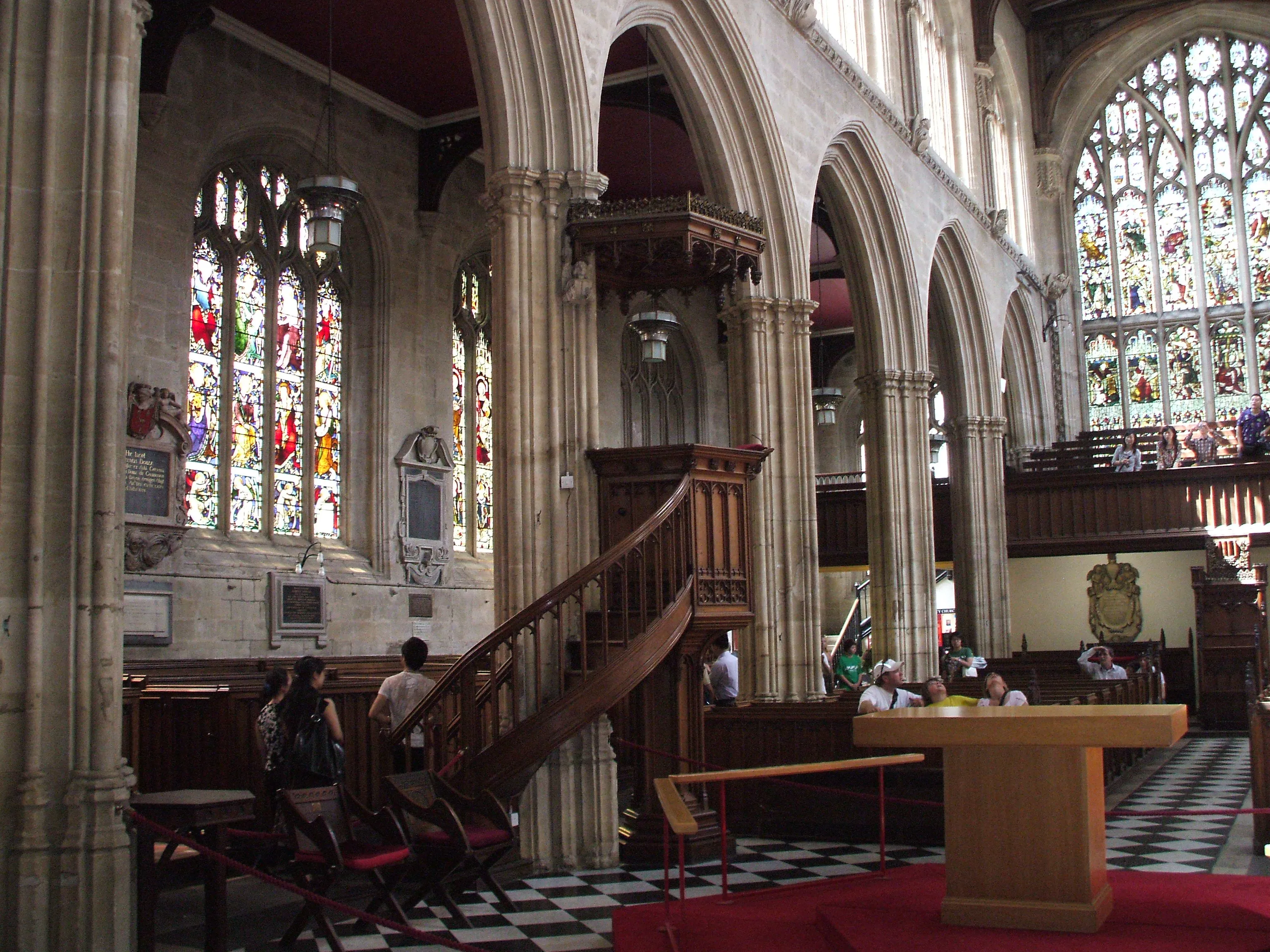 Interior de la University Church of St Mary the Virgin que muestra el púlpito de madera ornamentado con escalera, arcos de piedra góticos, vidrieras y suelo de tablero de ajedrez con visitantes sentados cerca.