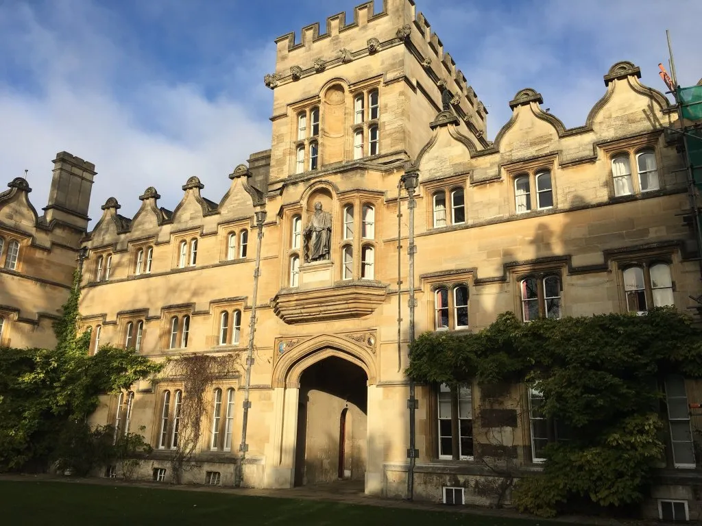 Stone facade of University College Oxford featuring a central arched gateway with a statue in a niche above, flanked by ivy-covered walls and multiple windows under a blue sky
