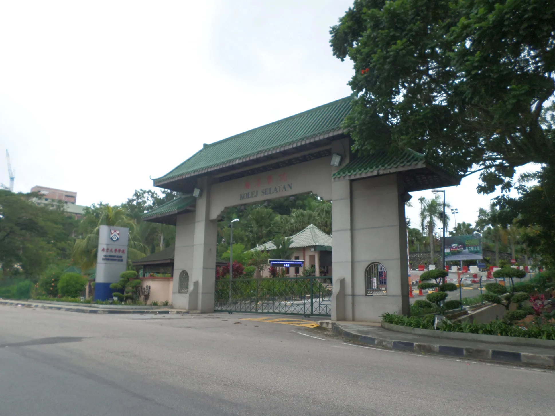 Entrance gate with green traditional tiled roof and University College signage, flanked by tropical trees and a paved road