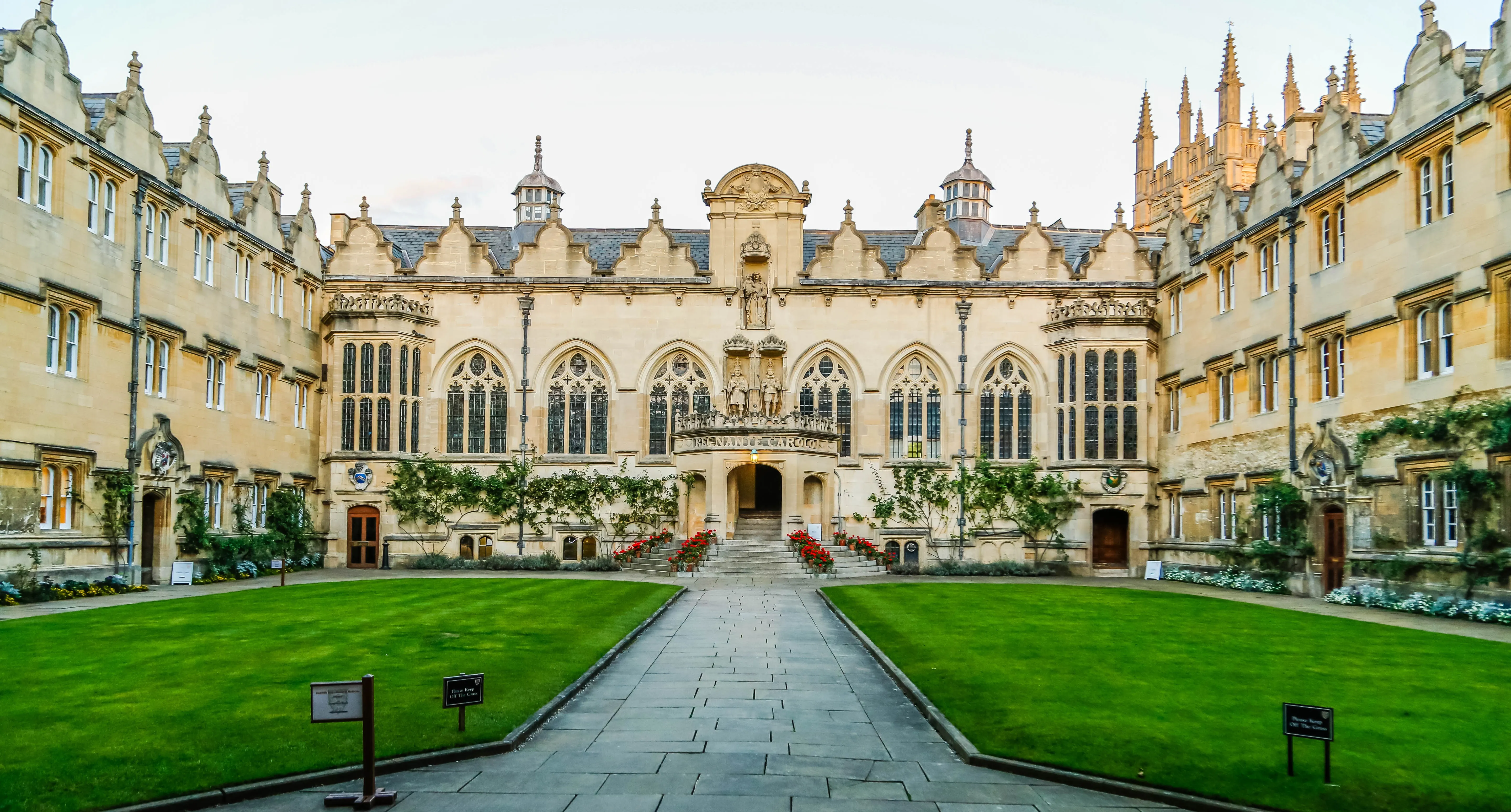 Stone courtyard with central pathway leading to a Gothic-style building facade, flanked by green lawns and ornate historic architecture