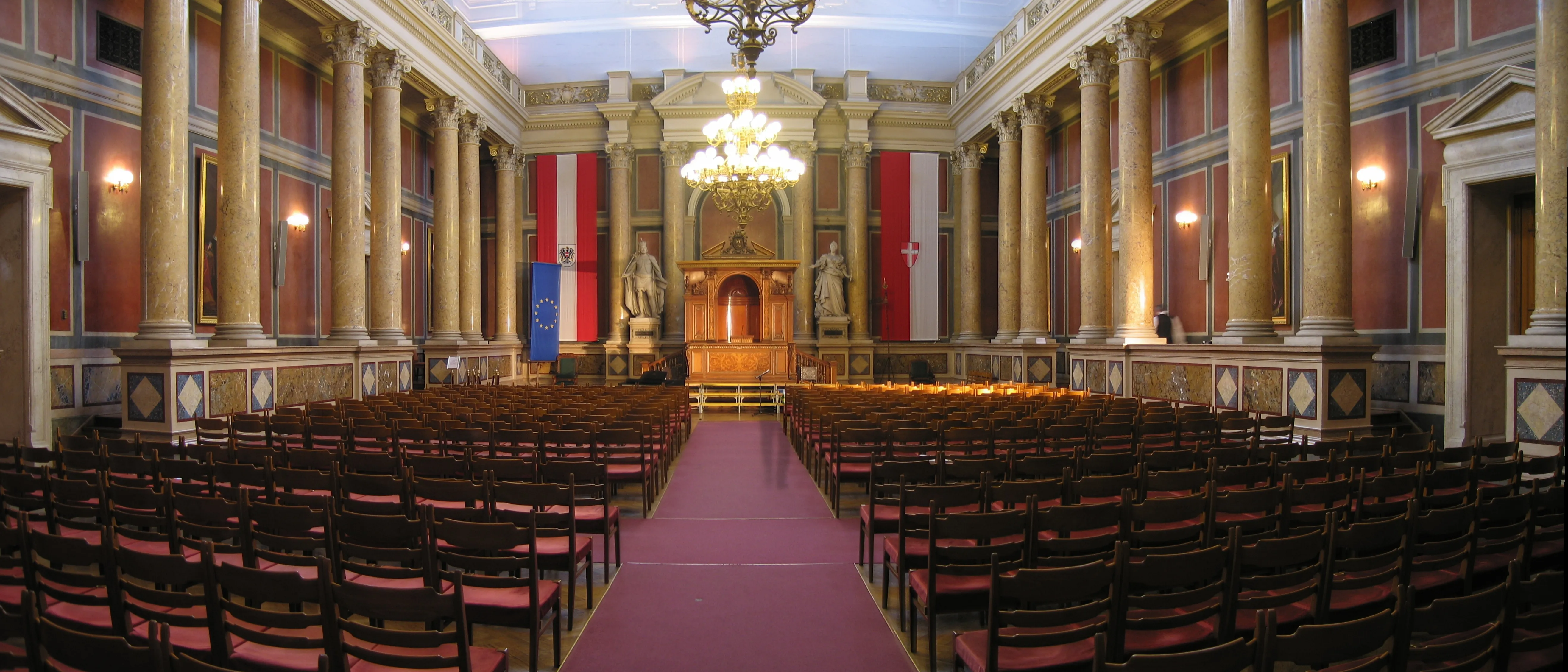 Gran salón con filas de sillas de madera, una alfombra roja, altas columnas y una lámpara de araña en la Universidad de Viena.